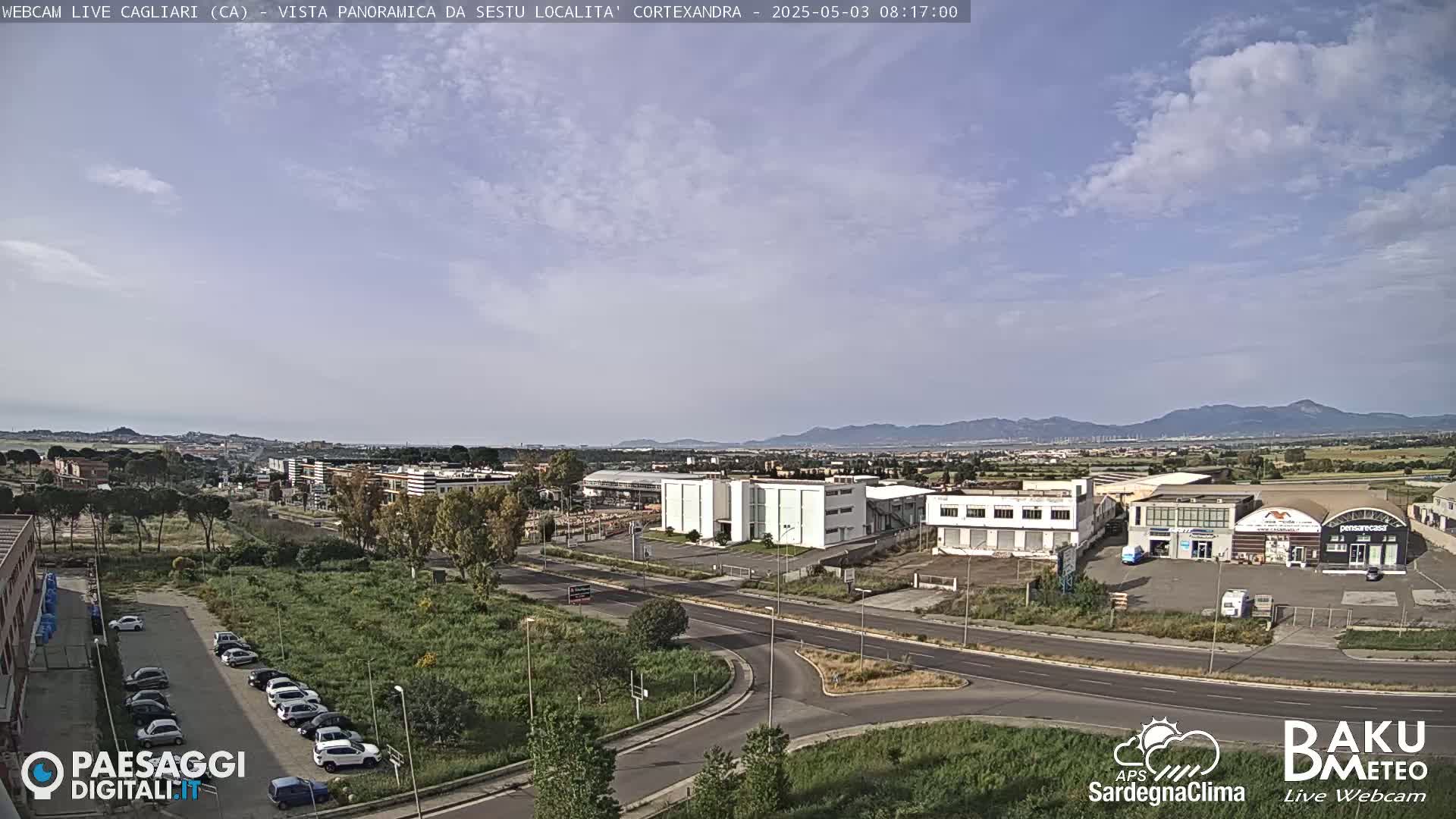 A partly cloudy day reveals a panoramic view of a city with industrial buildings and a parking lot filled with cars, all situated against a backdrop of distant mountains.