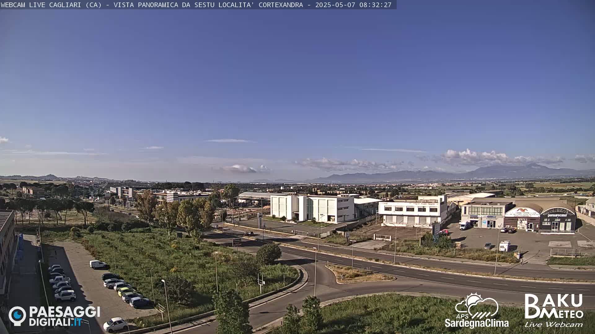 A mostly clear, sunny day reveals a panoramic view of industrial buildings and a road intersection, with mountains in the distance and a few scattered clouds in the sky.