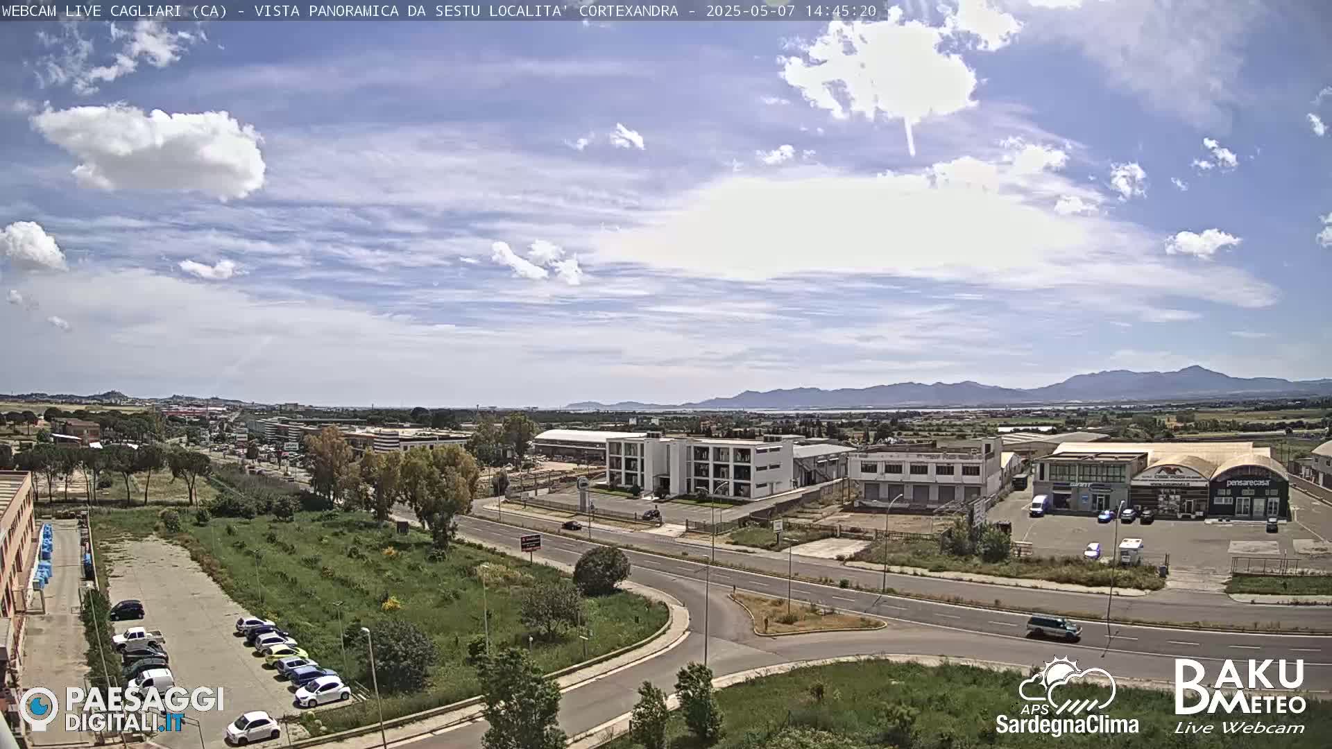 A partly cloudy day reveals a view of a city with industrial buildings, roads, and a parking lot filled with cars, all situated before a range of distant mountains.