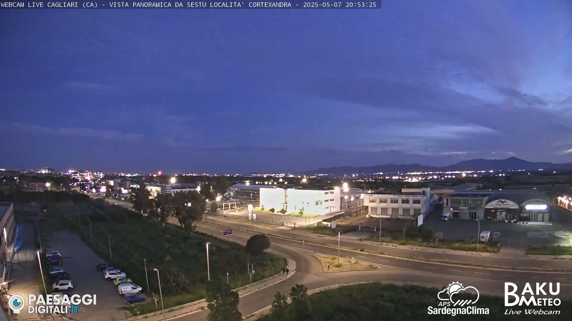 A twilight view of a city and surrounding landscape, showing roads, buildings, and parked cars under a mostly clear, dark blue sky.