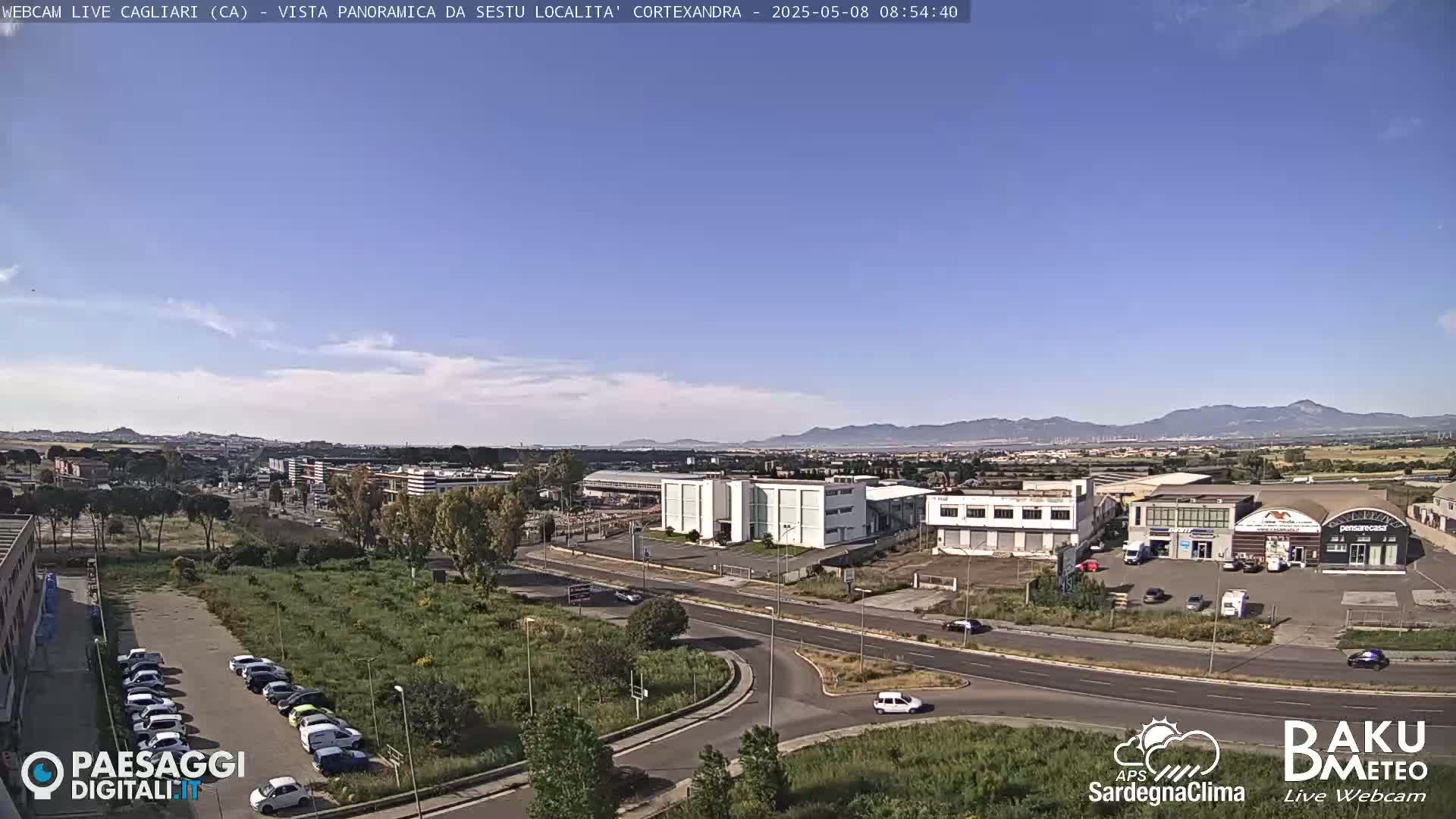 A mostly clear, sunny day overlooks a sprawling industrial area with buildings, roads, and a parking lot, and distant mountains in the background.