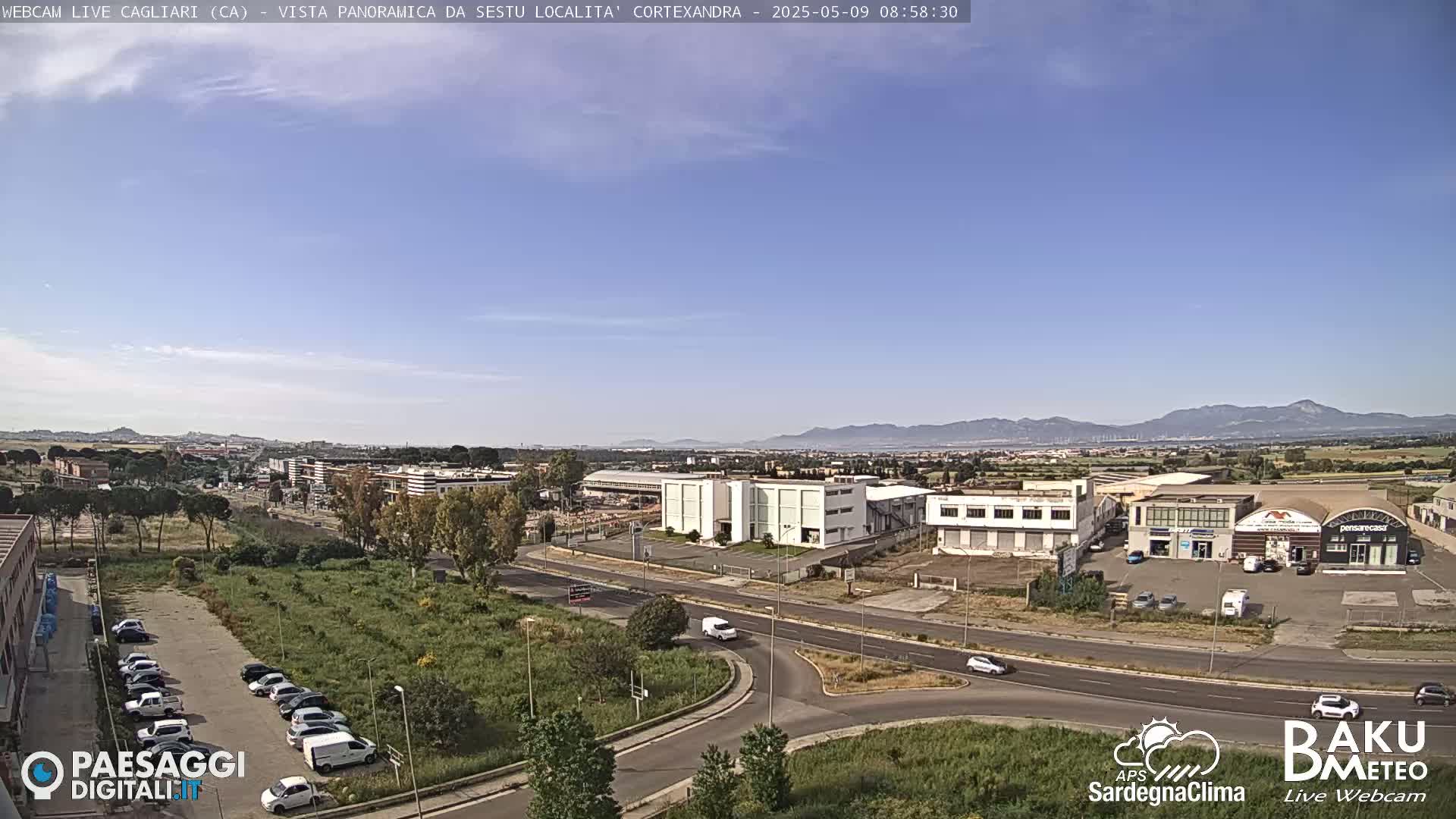A panoramic view of a city under a clear blue sky shows a roadway intersection with various buildings and parked cars, surrounded by green vegetation and distant mountains.