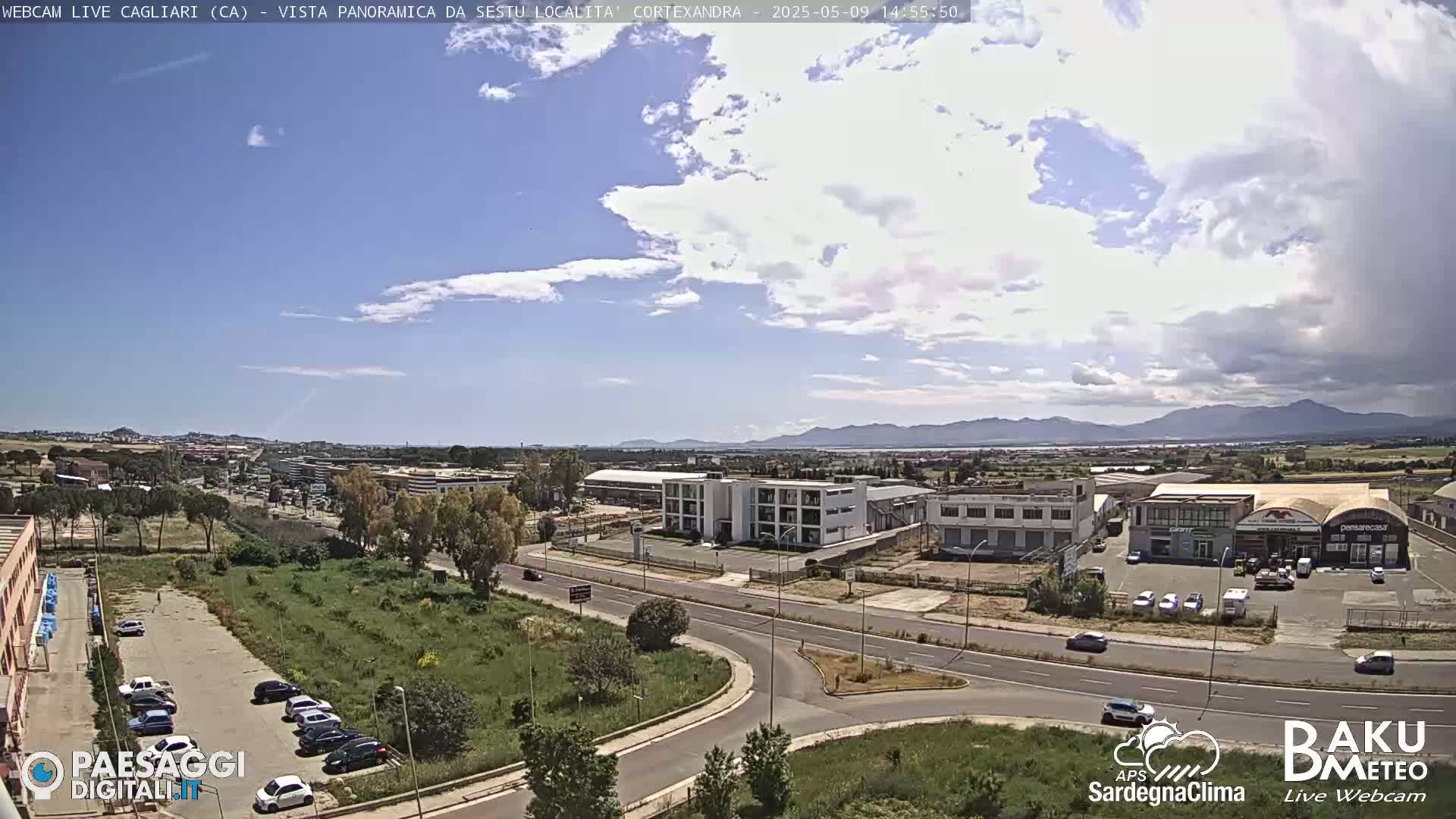 A partly cloudy day reveals a wide shot of a city road intersection, surrounded by buildings and parking lots, with distant mountains under a mostly clear blue sky.