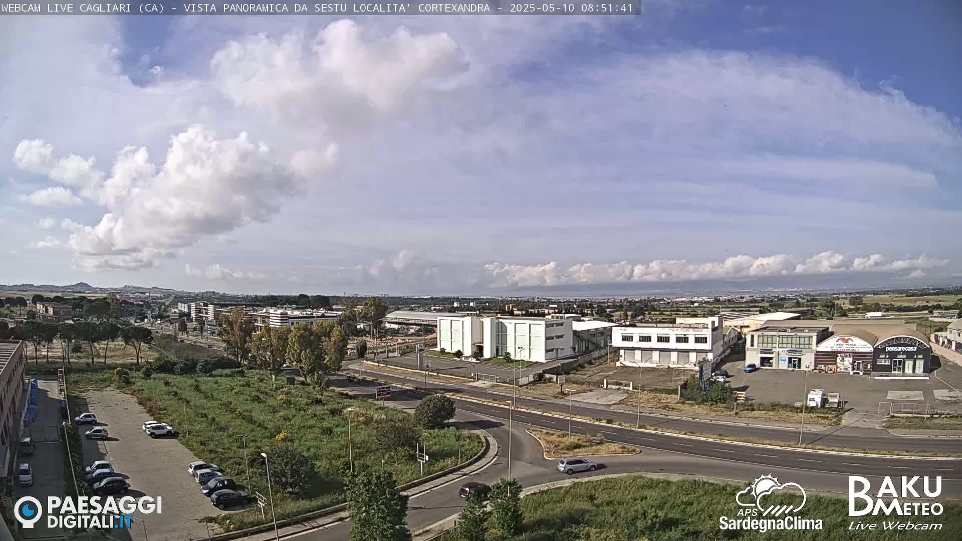 A partly cloudy day reveals a wide shot of a suburban area with roads, buildings, and parked cars, overlooking a distant body of water.