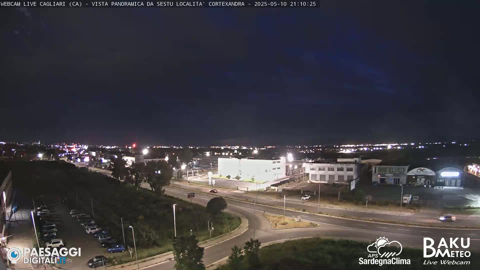 A nighttime, panoramic view of a city's outskirts, featuring roads, buildings, and a few parked cars under a clear, dark sky.