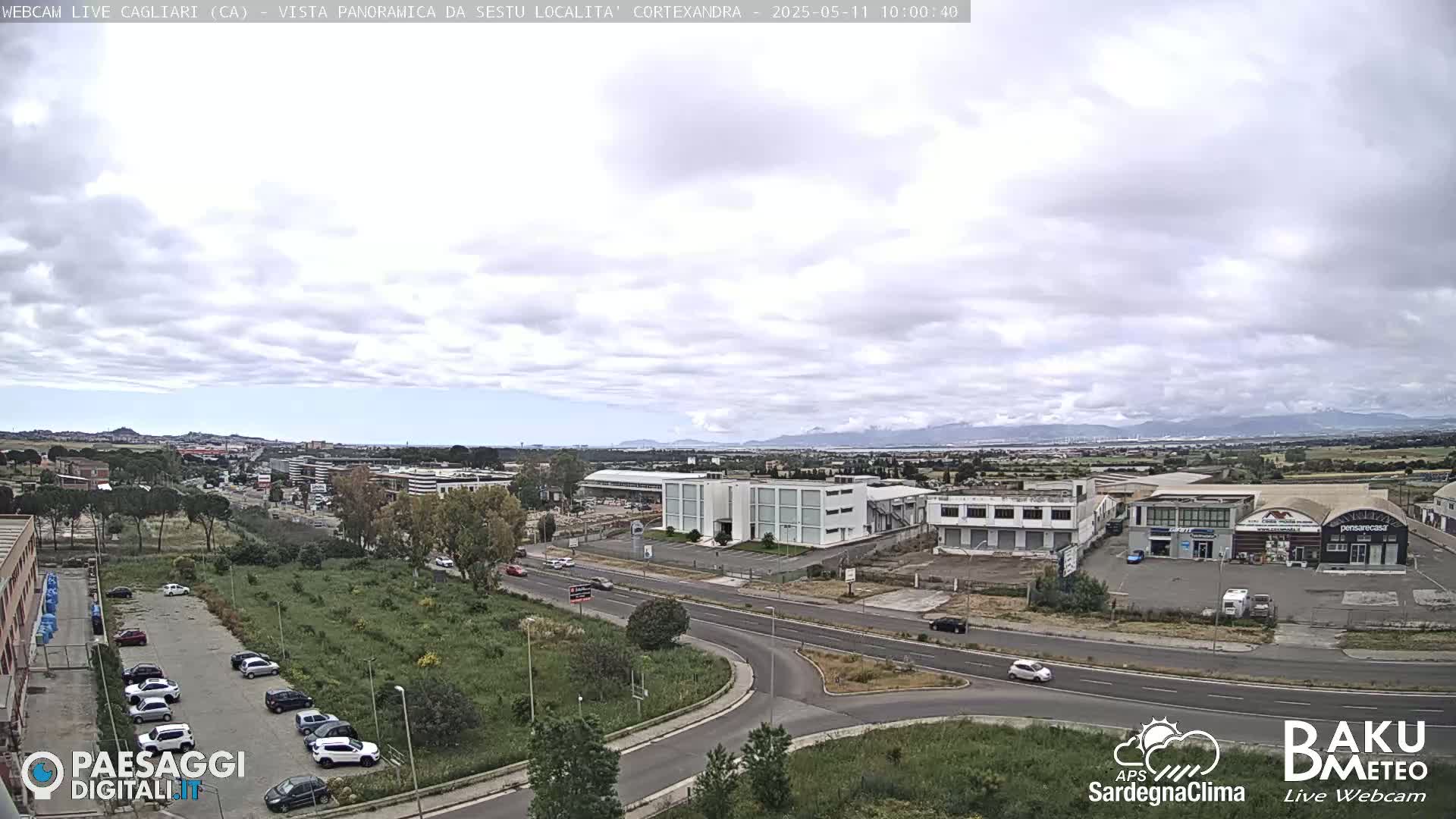A mostly cloudy day reveals a panoramic view of a city with roads, buildings, and parked cars, mountains visible in the distance.
