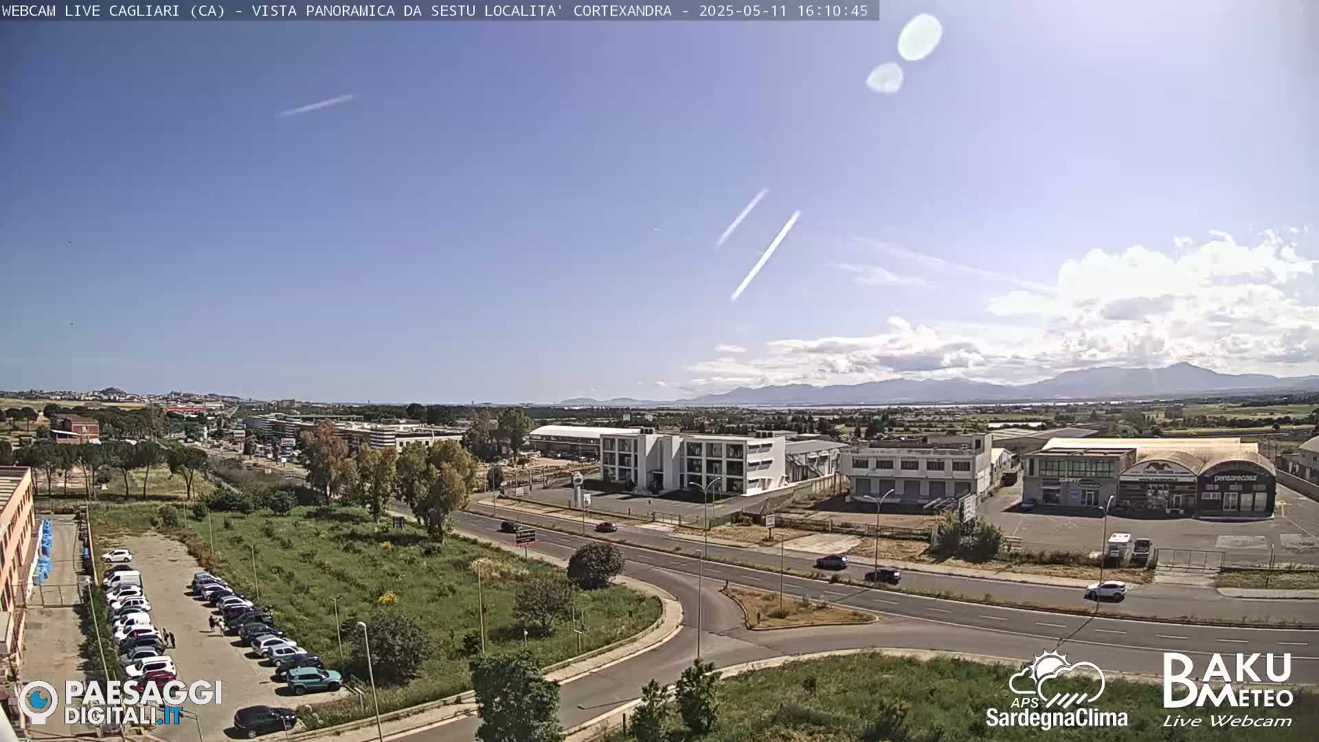 A panoramic view of a city with buildings, roads, and parked cars under a mostly clear sky with a few clouds and contrails.