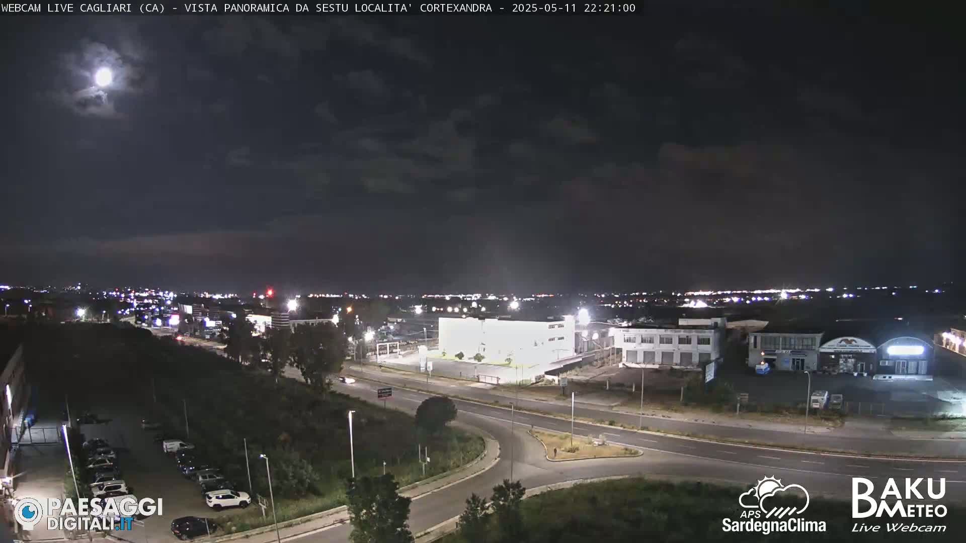 A nighttime, panoramic view of a city with a bright moon in the sky, showing roads, buildings, and sparse vegetation.