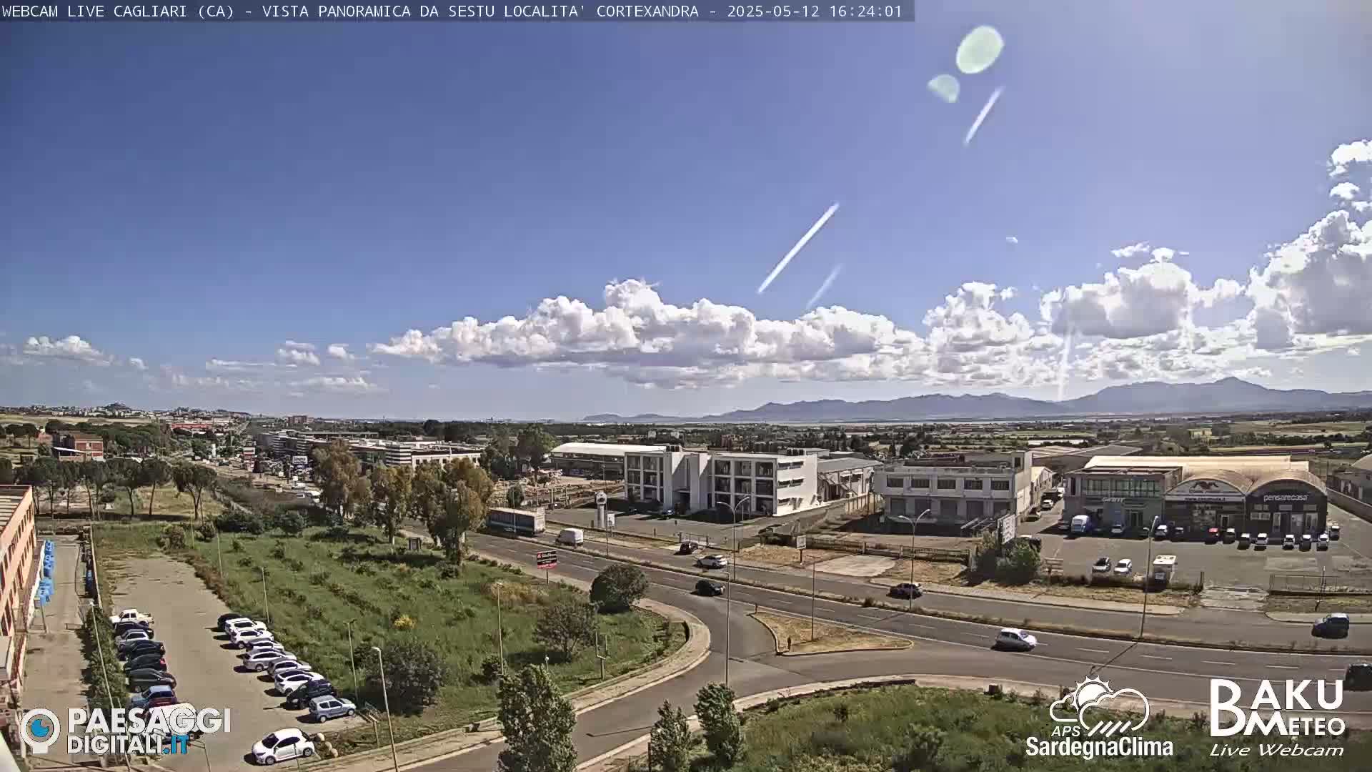 A partly cloudy daytime view of a road intersection, industrial buildings, and parking lots, with distant mountains visible under a mostly clear sky.