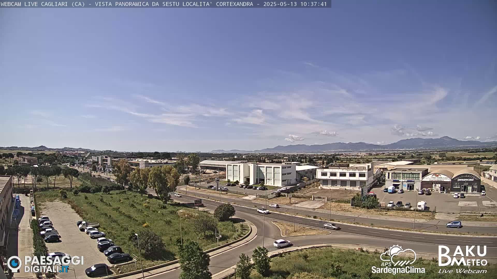 A panoramic view of a city with light traffic under a mostly clear, sunny sky, featuring distant mountains on the horizon.