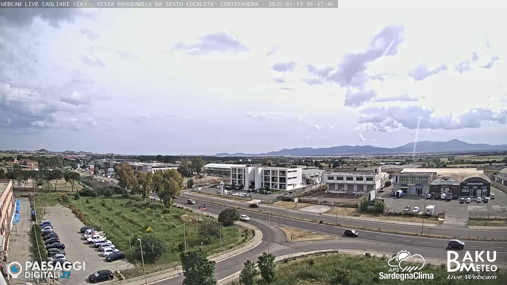 A partly cloudy day reveals a wide, aerial view of a suburban area with roads, buildings, and parked cars, set against a backdrop of distant mountains.
