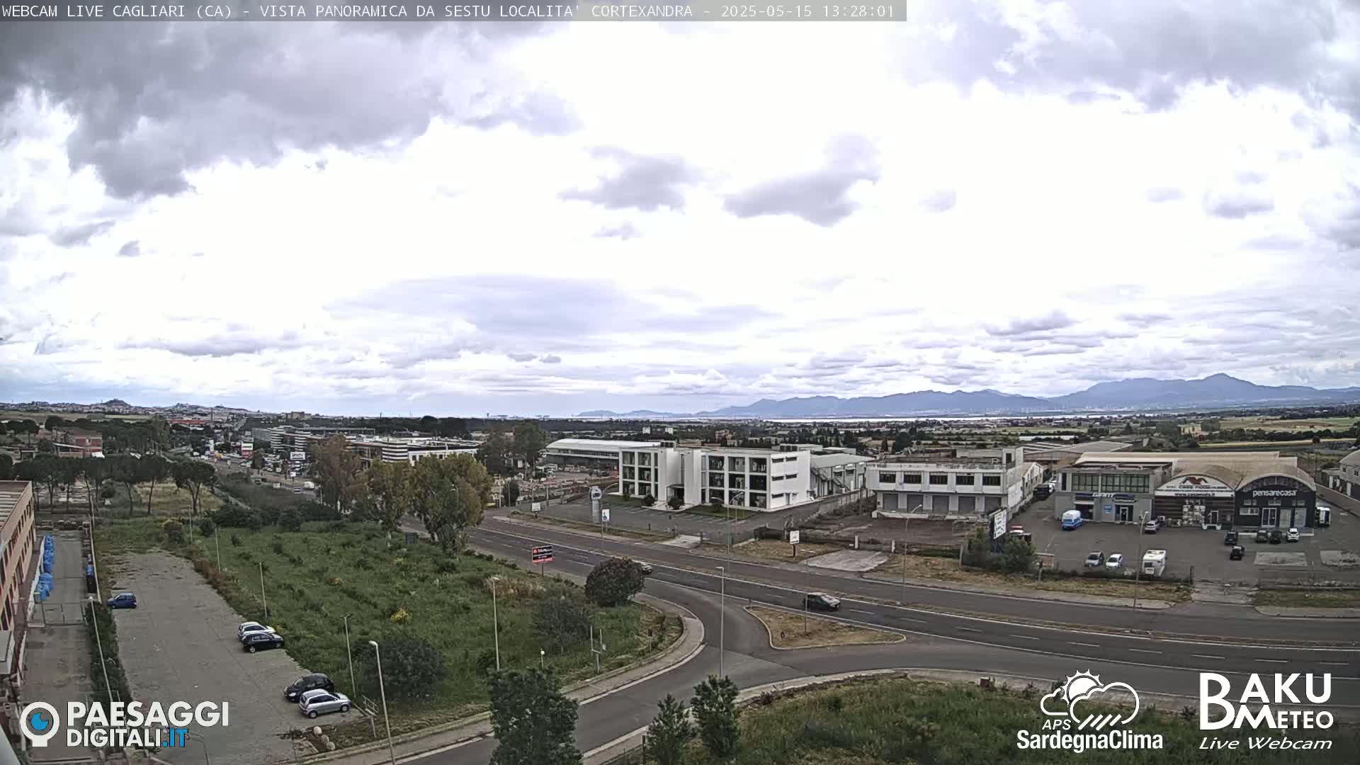 A partly cloudy day reveals a wide shot of a roadway intersection, industrial buildings, and distant mountains, with several cars visible in parking lots and on the road.
