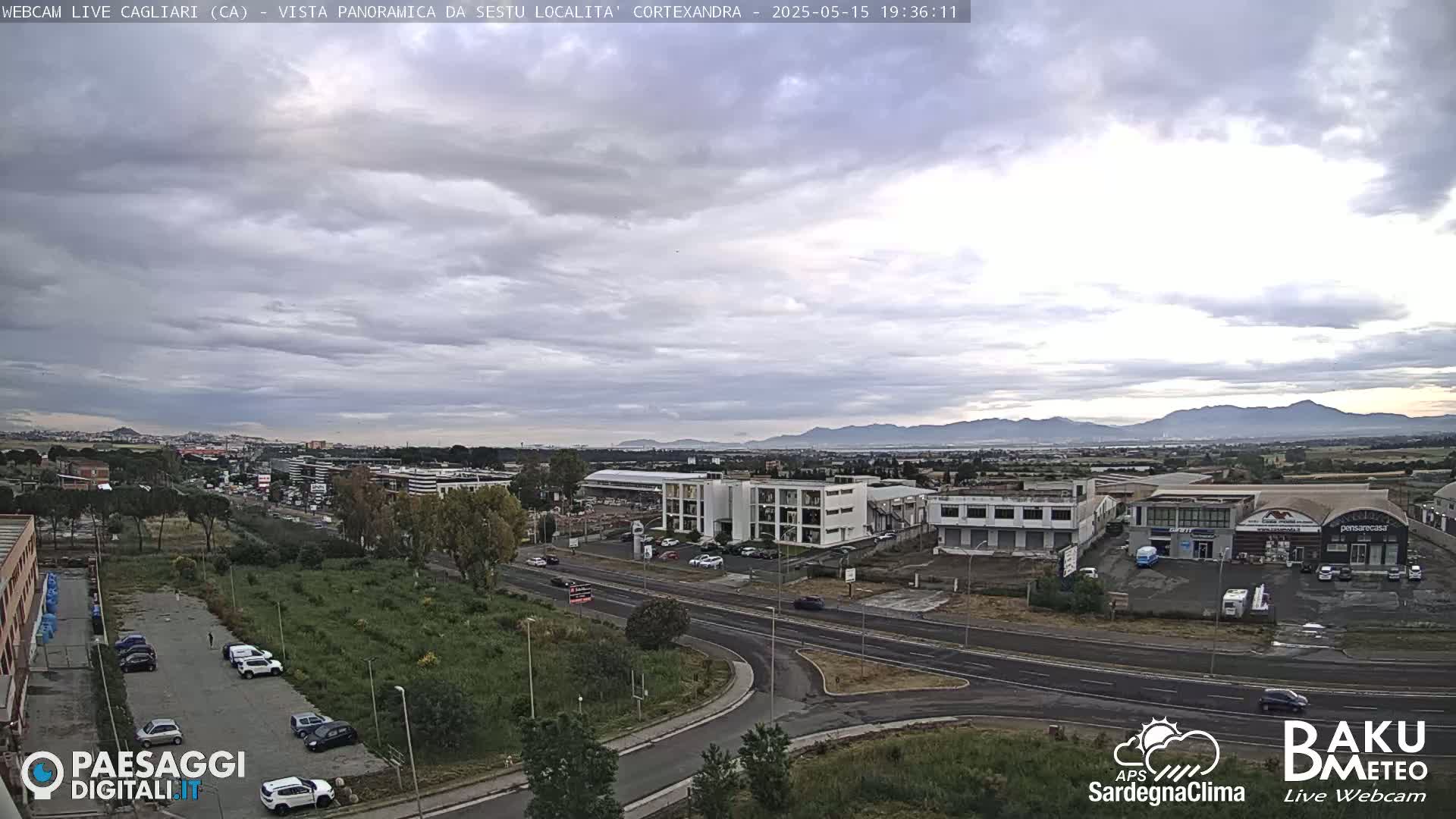 A panoramic view of a city under a mostly cloudy sky shows a highway intersection, several buildings, and distant mountains.