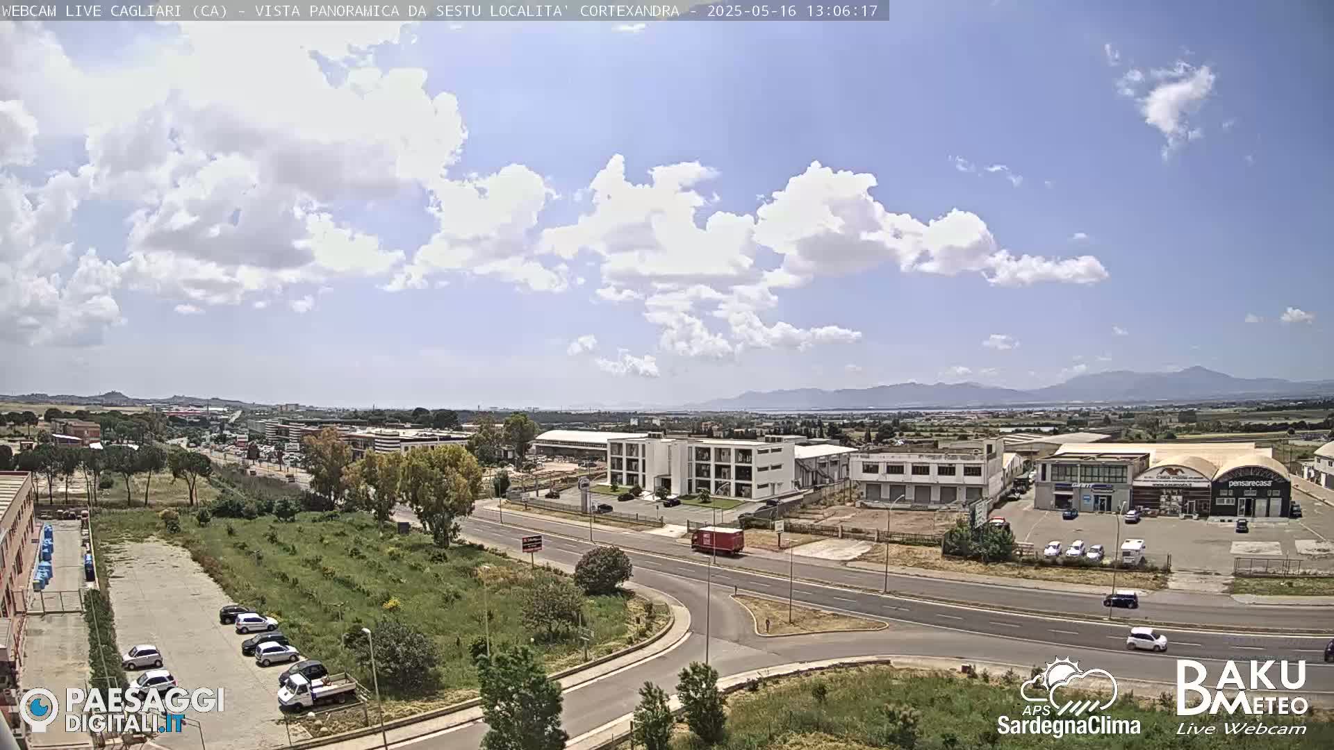 A partly sunny day shows a view of a highway intersection, industrial buildings, and parked cars, with mountains visible in the distance.