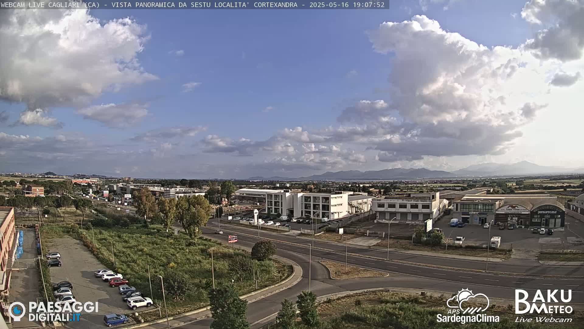 A partly cloudy afternoon scene shows a wide shot of a road intersection and surrounding buildings, with mountains visible in the distance.