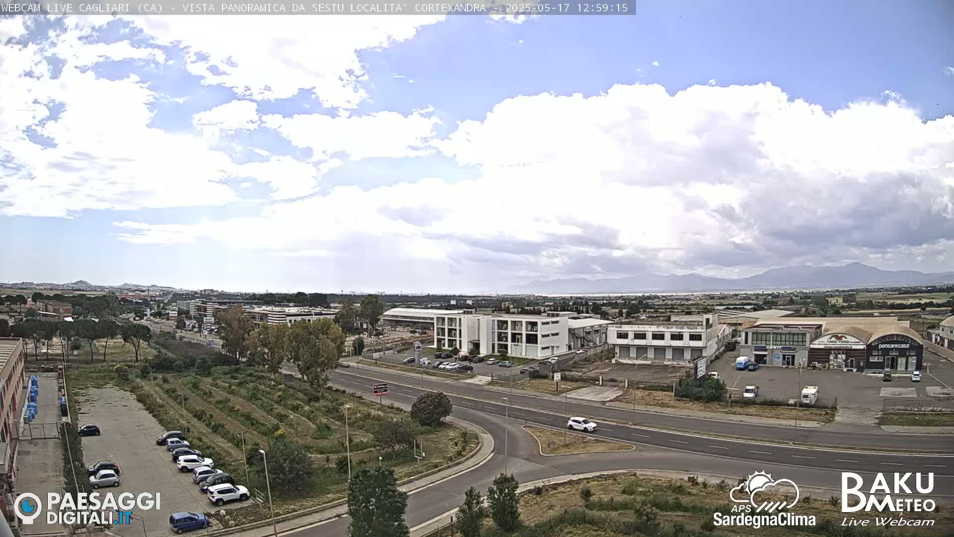 A partly cloudy day reveals a wide shot of a city landscape with industrial buildings, roads, and a parking lot filled with cars, all situated before a distant mountain range.