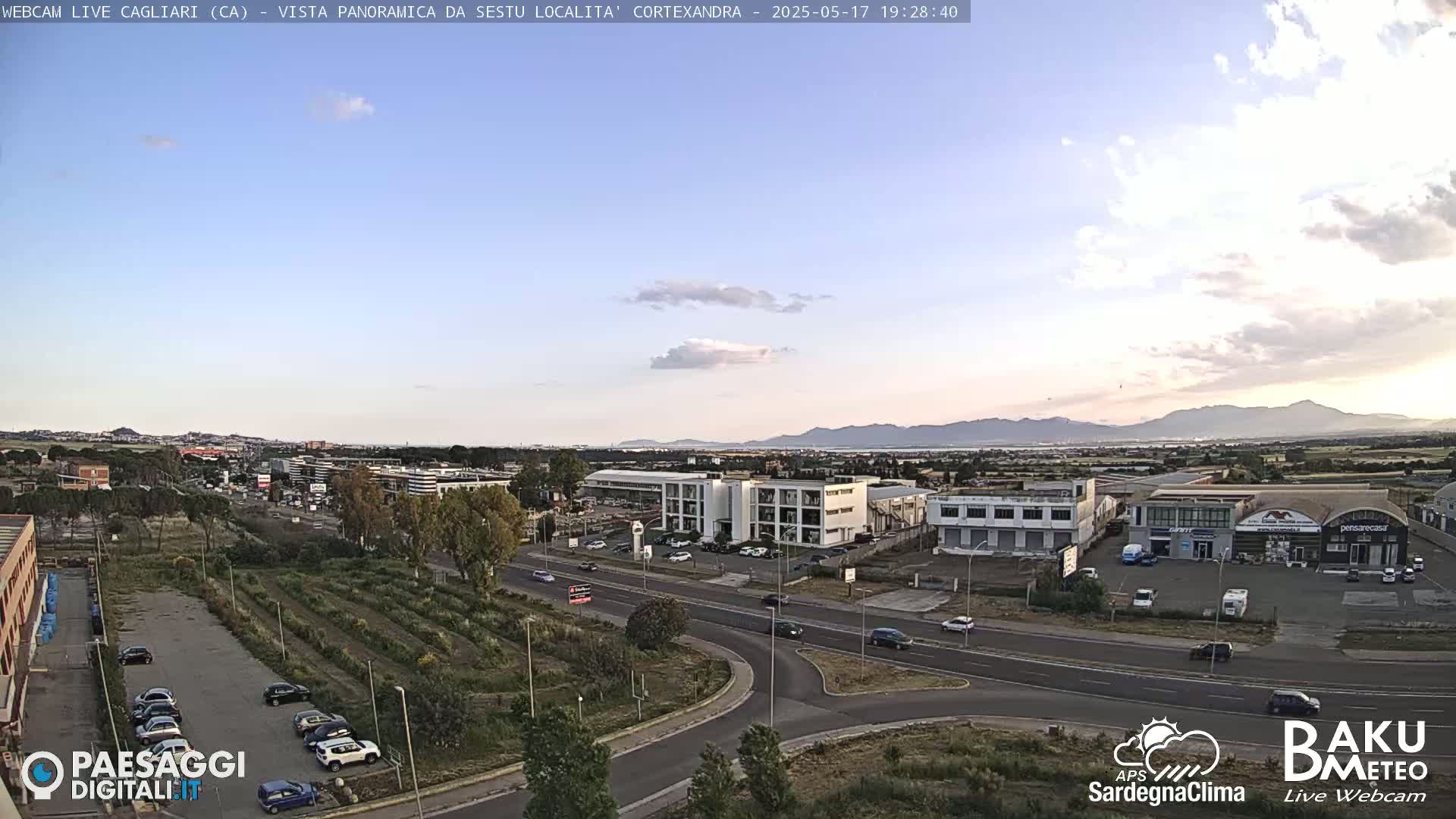 A panoramic view of a city under a partly cloudy evening sky shows a road intersection with cars, buildings, and a field of vegetation in the foreground, and mountains in the distance.