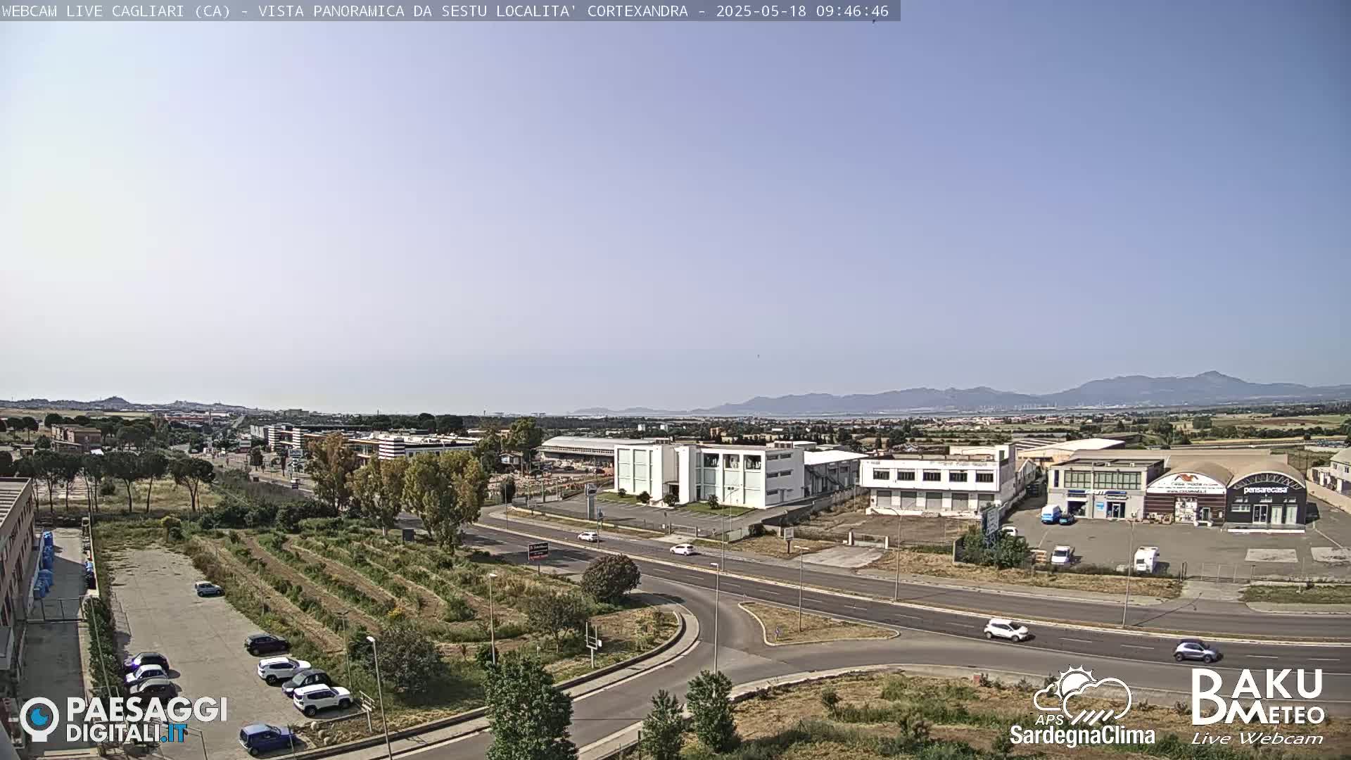 A clear, sunny day reveals a panoramic view of an industrial area with buildings, roads, and sparse vegetation, extending to distant mountains under a pale blue sky.