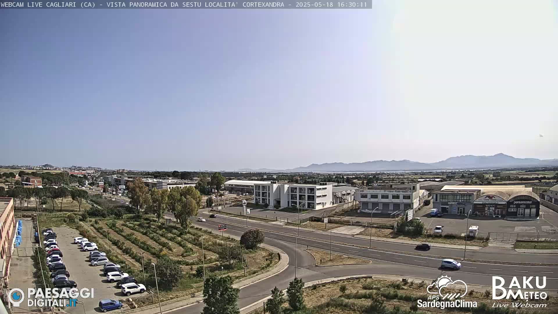 A sunny, clear day overlooking a suburban area with roads, buildings, a parking lot full of cars, and a cultivated field, all set against a backdrop of distant mountains.