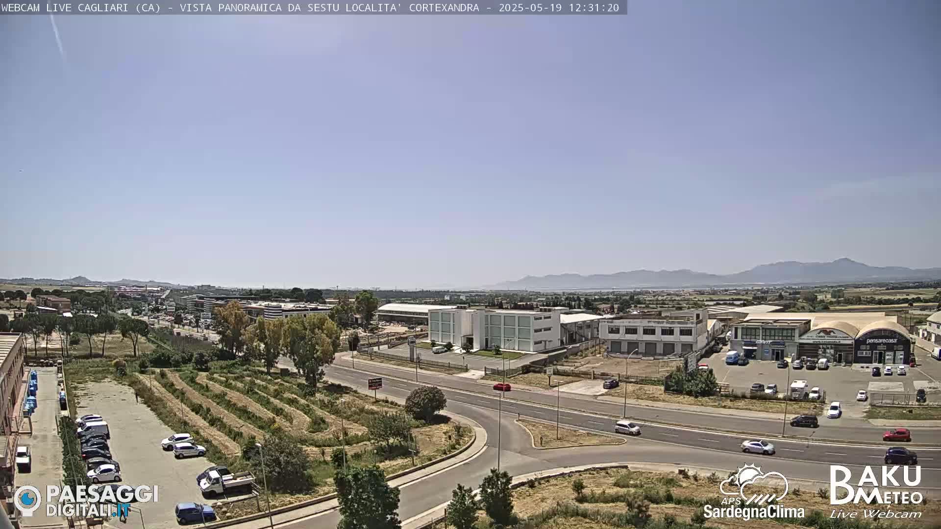 A sunny, clear day reveals a panoramic view of an industrial area, with roads, buildings, and sparse vegetation, backed by distant mountains.