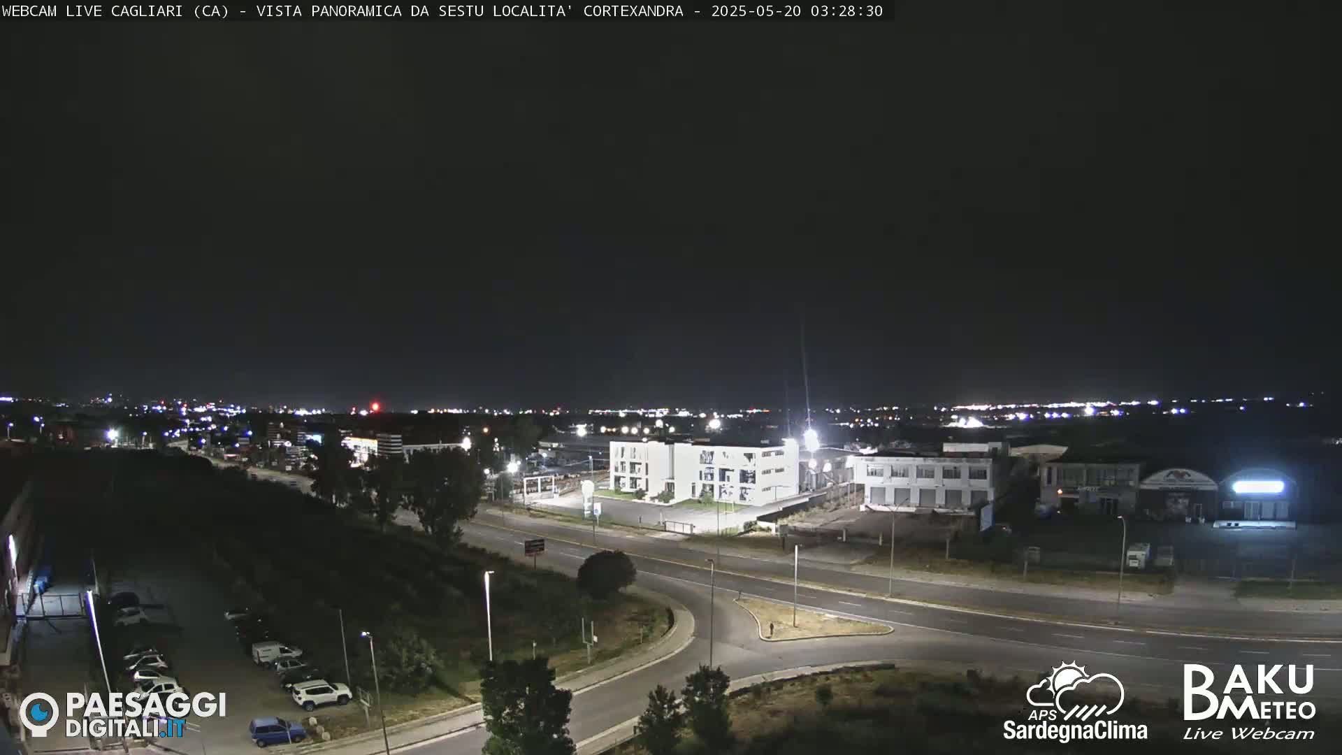 A nighttime, panoramic view of a city's outskirts, showing roads, buildings, and sparse vegetation under a clear, dark sky.