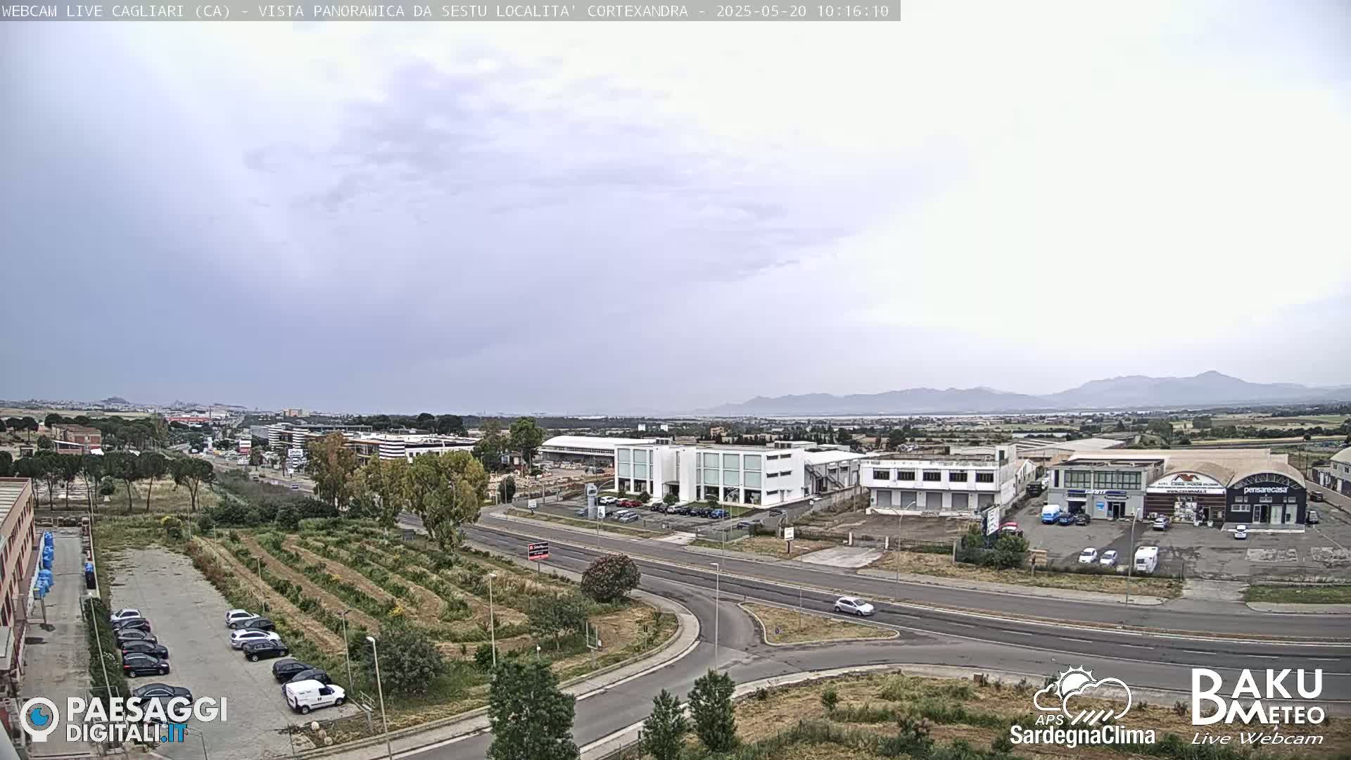 A panoramic view of a city with overcast skies shows a road intersection, numerous buildings, and a parking lot with cars, along with a cultivated area.