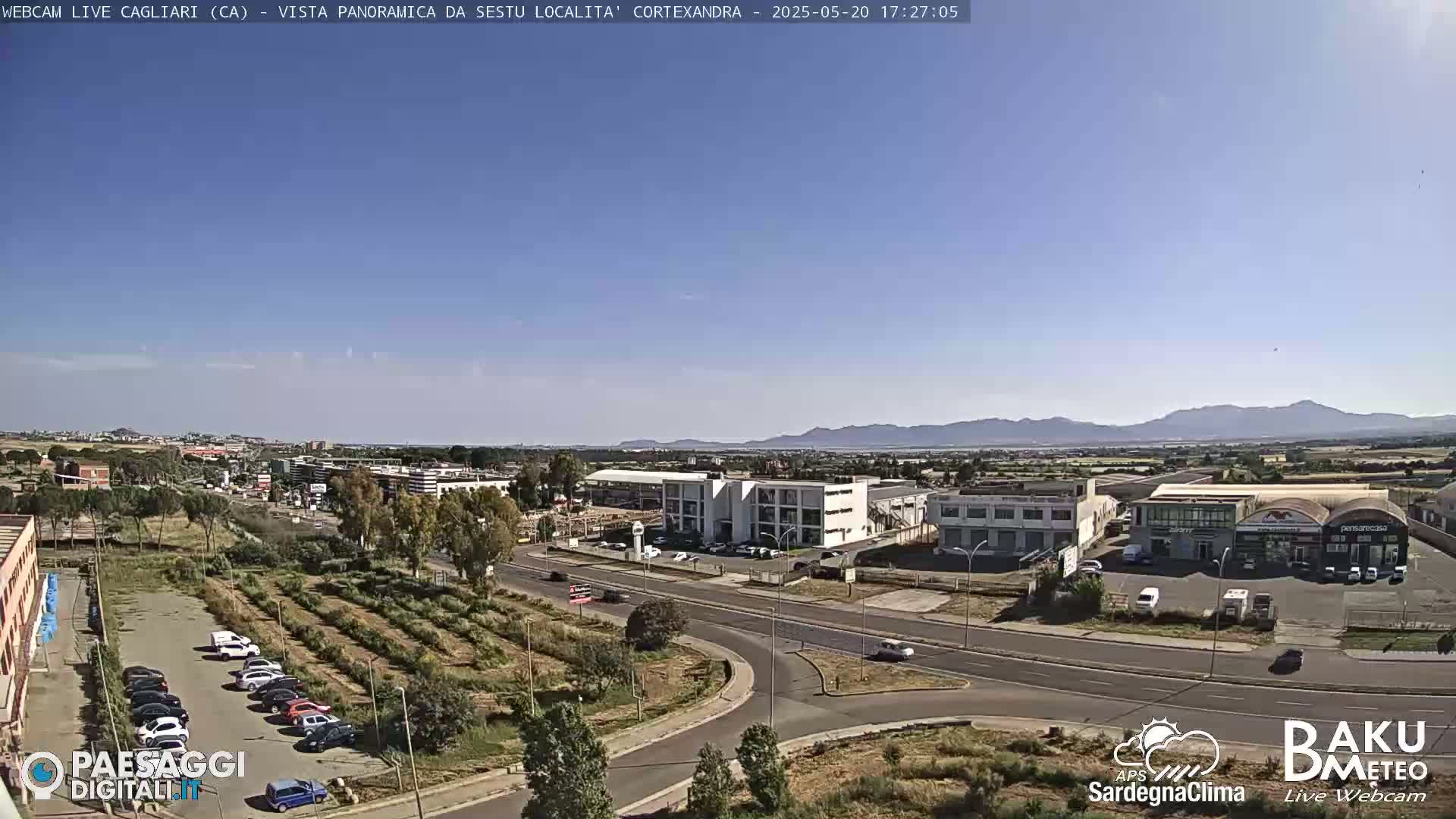 A clear, sunny day reveals a panoramic view of a city with industrial buildings, roads, and a parking lot filled with cars, all situated against a backdrop of distant mountains.