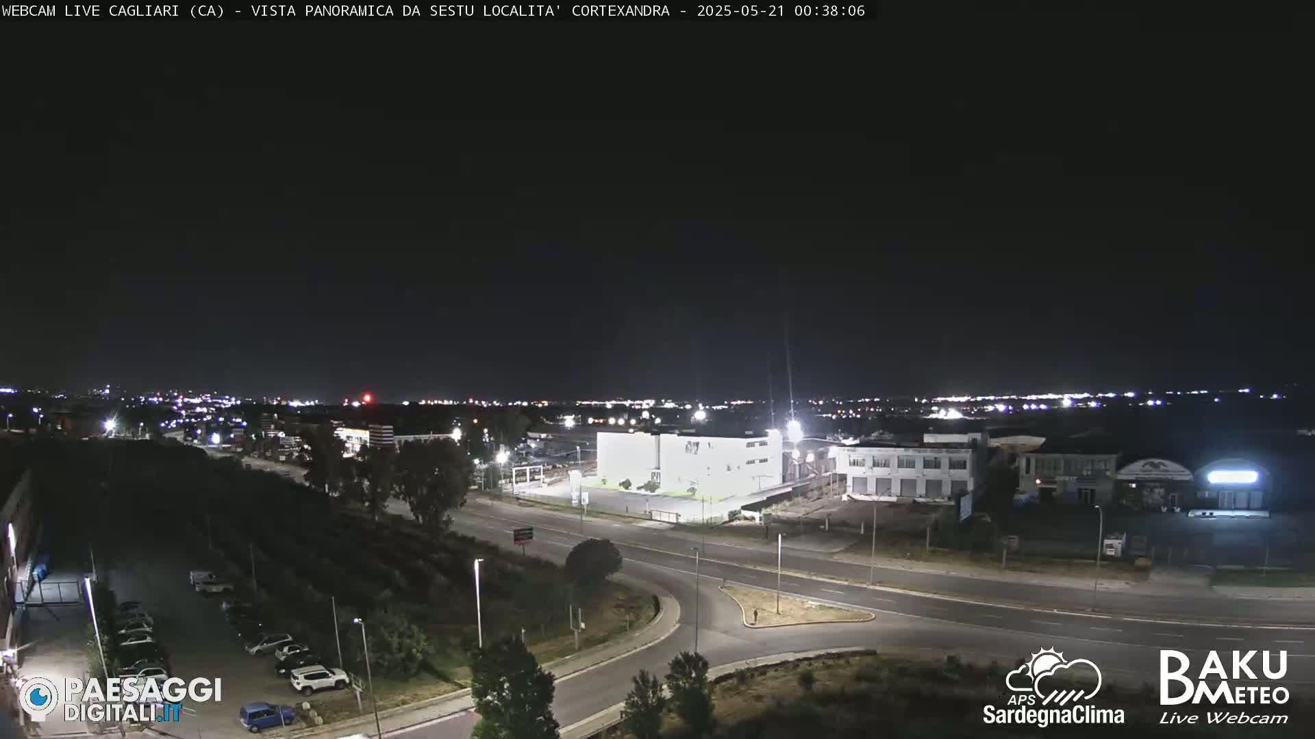 A nighttime, clear-sky view of a road intersection and industrial area, with city lights visible in the distance.