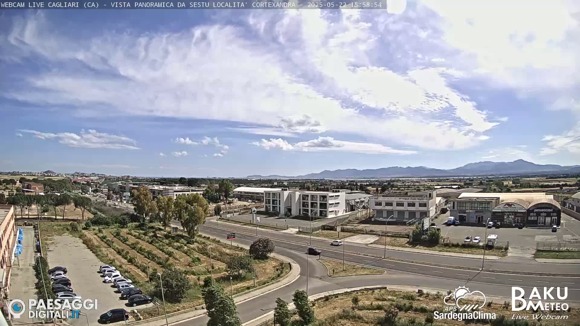 A partly cloudy day reveals a panoramic view of a city, featuring roads, buildings, and a parking lot with cars, all set against a backdrop of distant mountains.