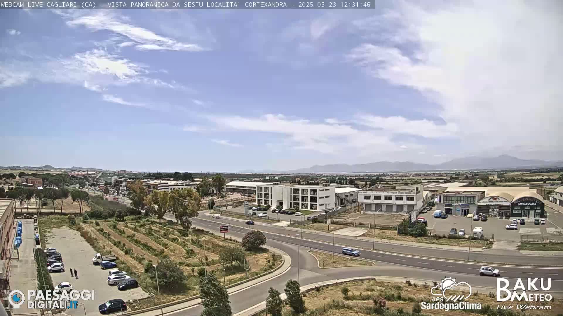 A mostly sunny day reveals a view of a suburban area with roads, buildings, and sparse vegetation, mountains visible in the distance.