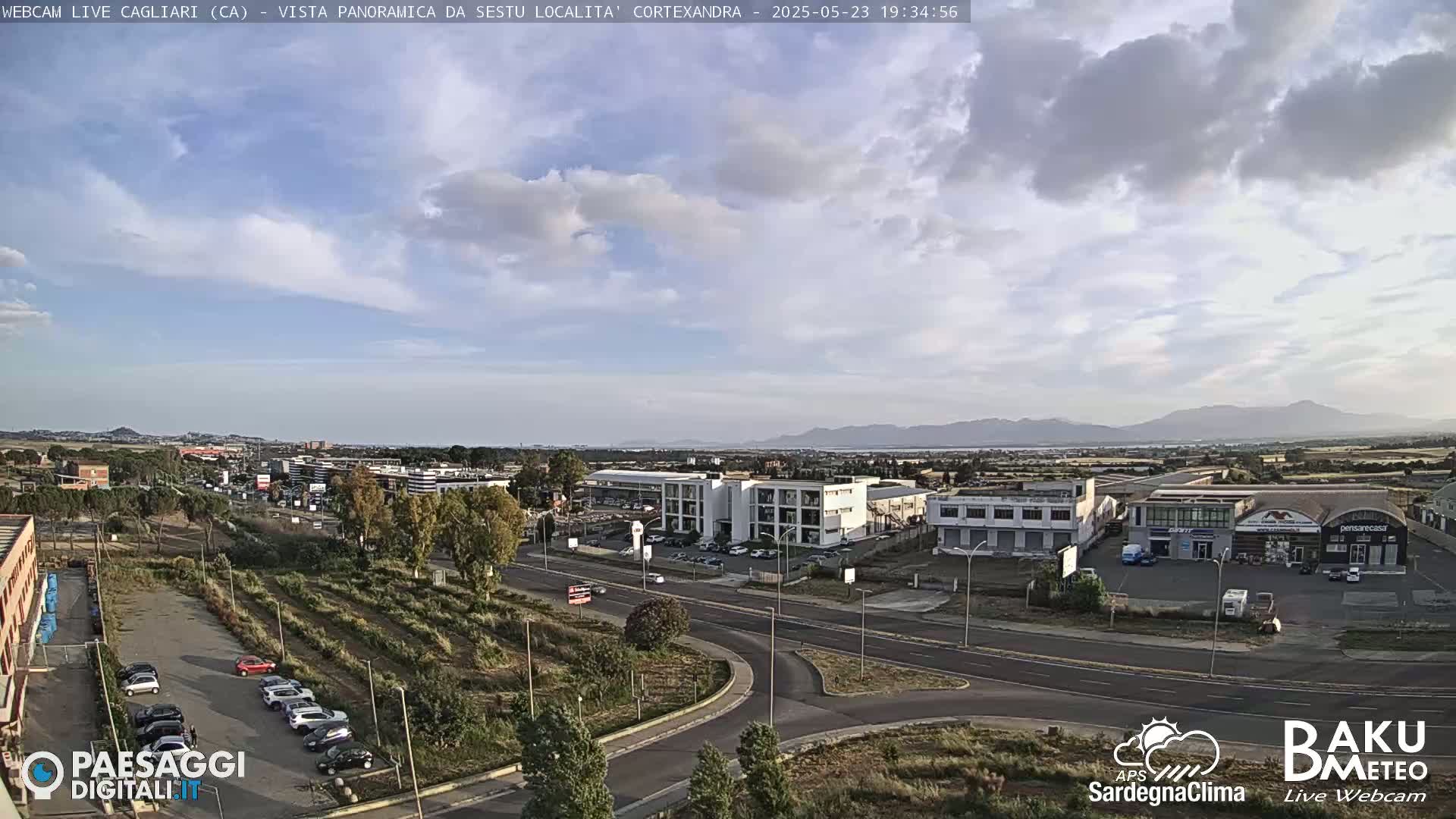 A mostly sunny, partly cloudy view of a city with buildings, roads, and cars, mountains visible in the distance.