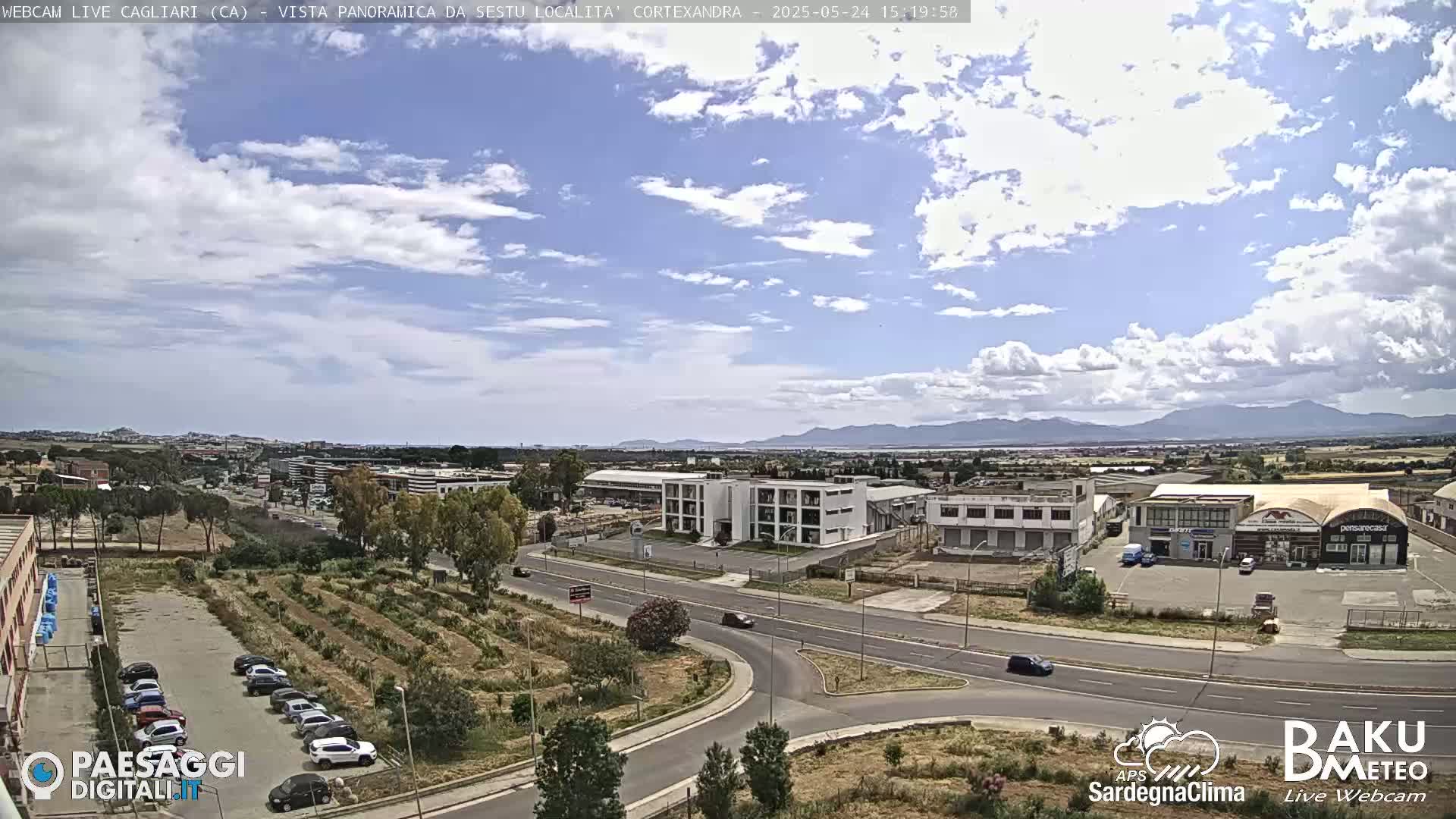 A partly cloudy day reveals a wide shot of a suburban area with roads, buildings, and a parking lot, all situated before a mountain range in the distance.