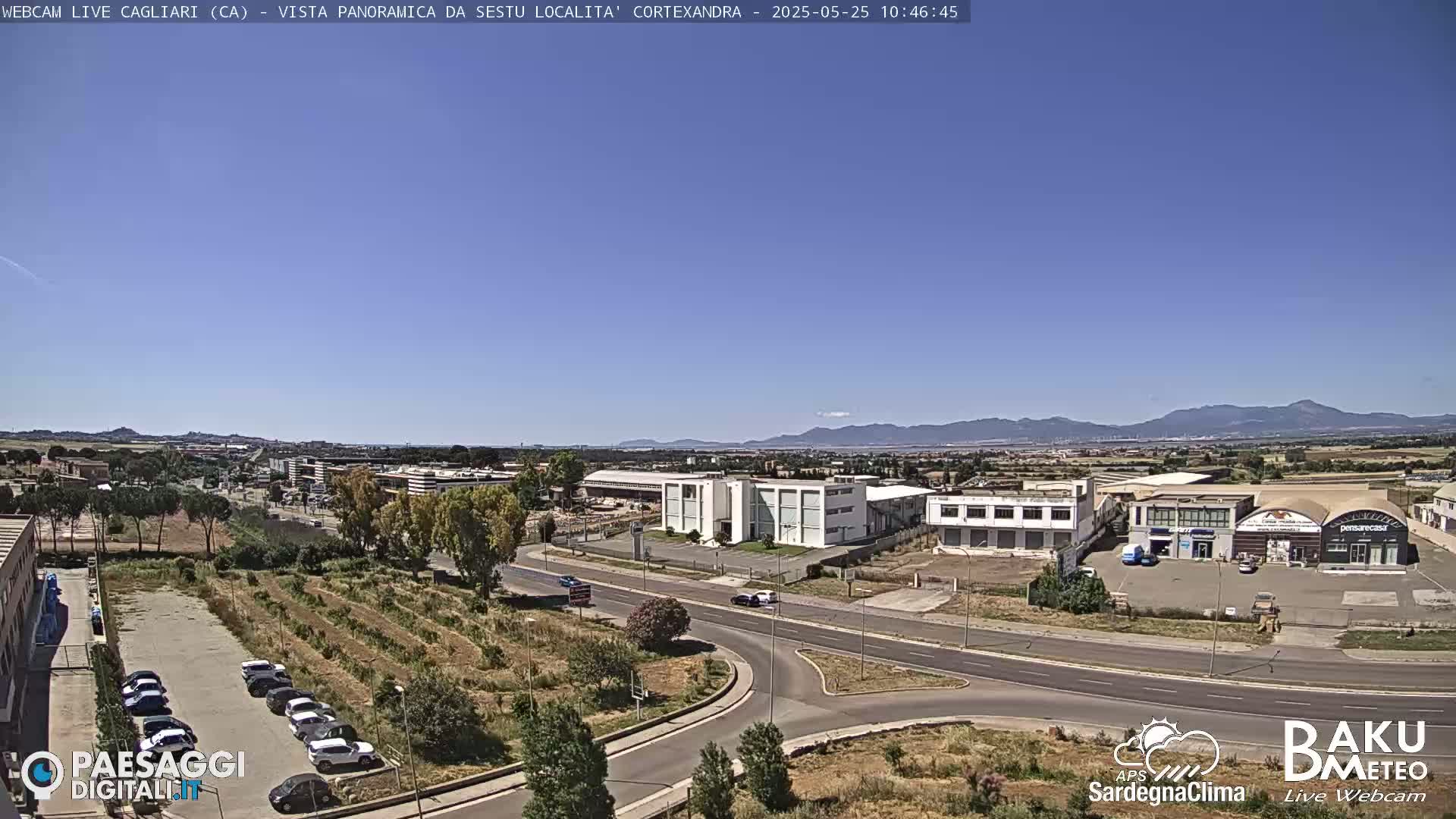 A clear, sunny day reveals a panoramic view of industrial buildings, roads, and a parking lot, all situated before a distant mountain range.