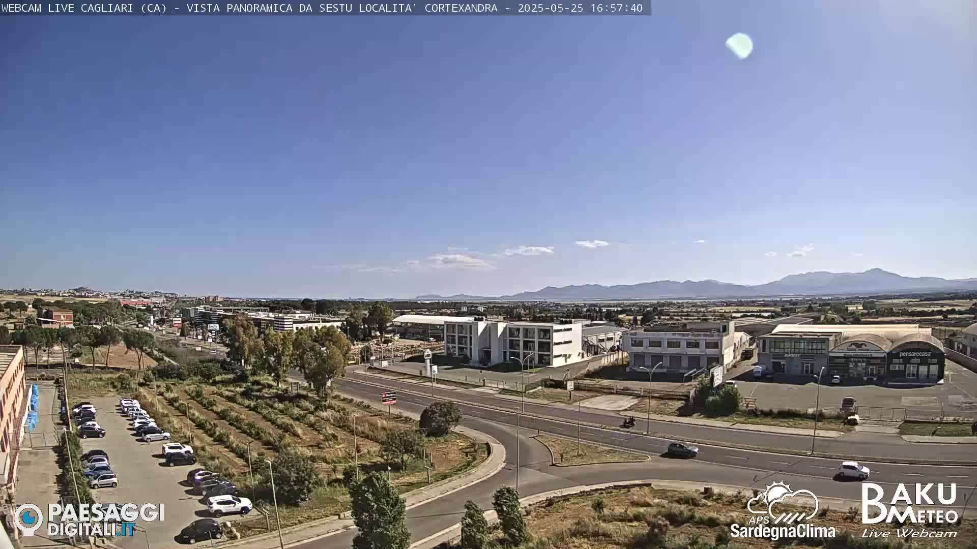 A clear, sunny day reveals a panoramic view of a city, including a road intersection, industrial buildings, a parking lot, and a distant mountain range.