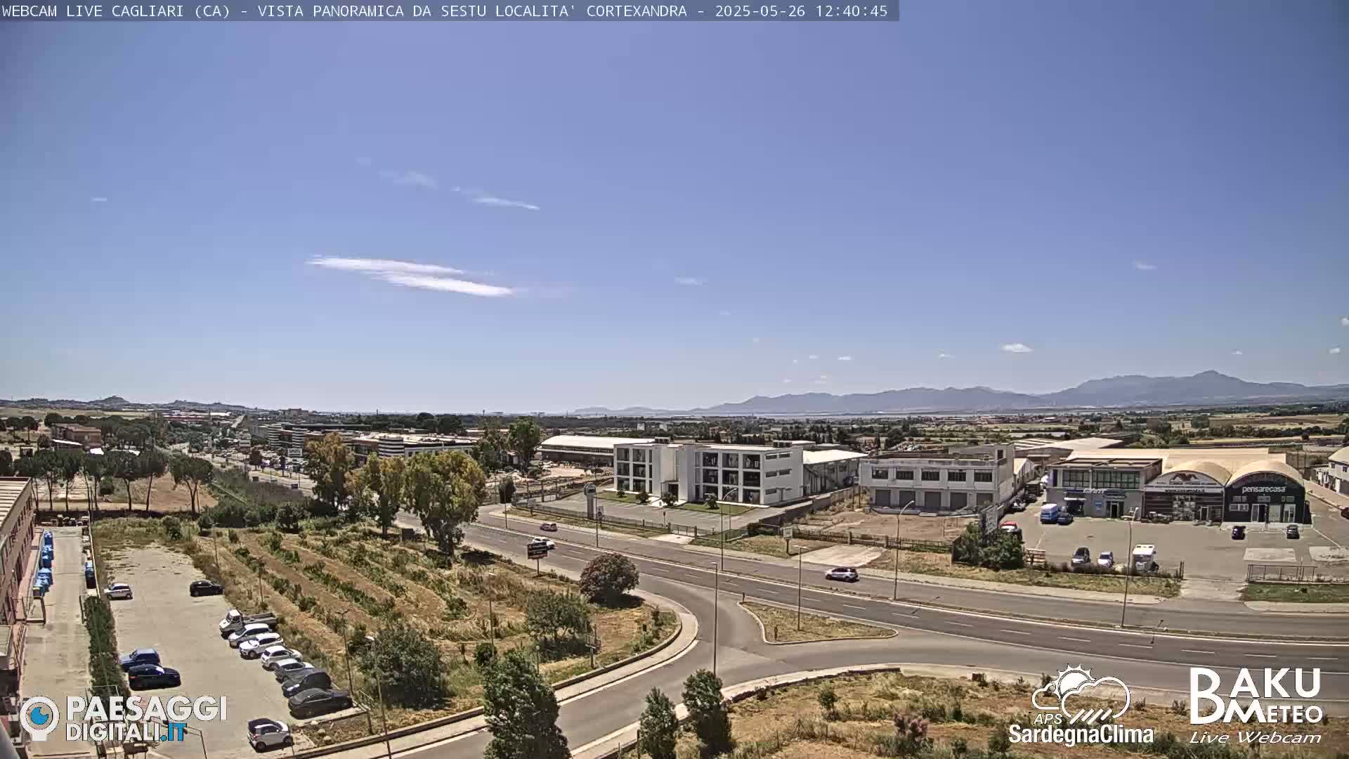 A sunny, clear day overlooking a city with industrial buildings and a roadway intersection, mountains visible in the distance.