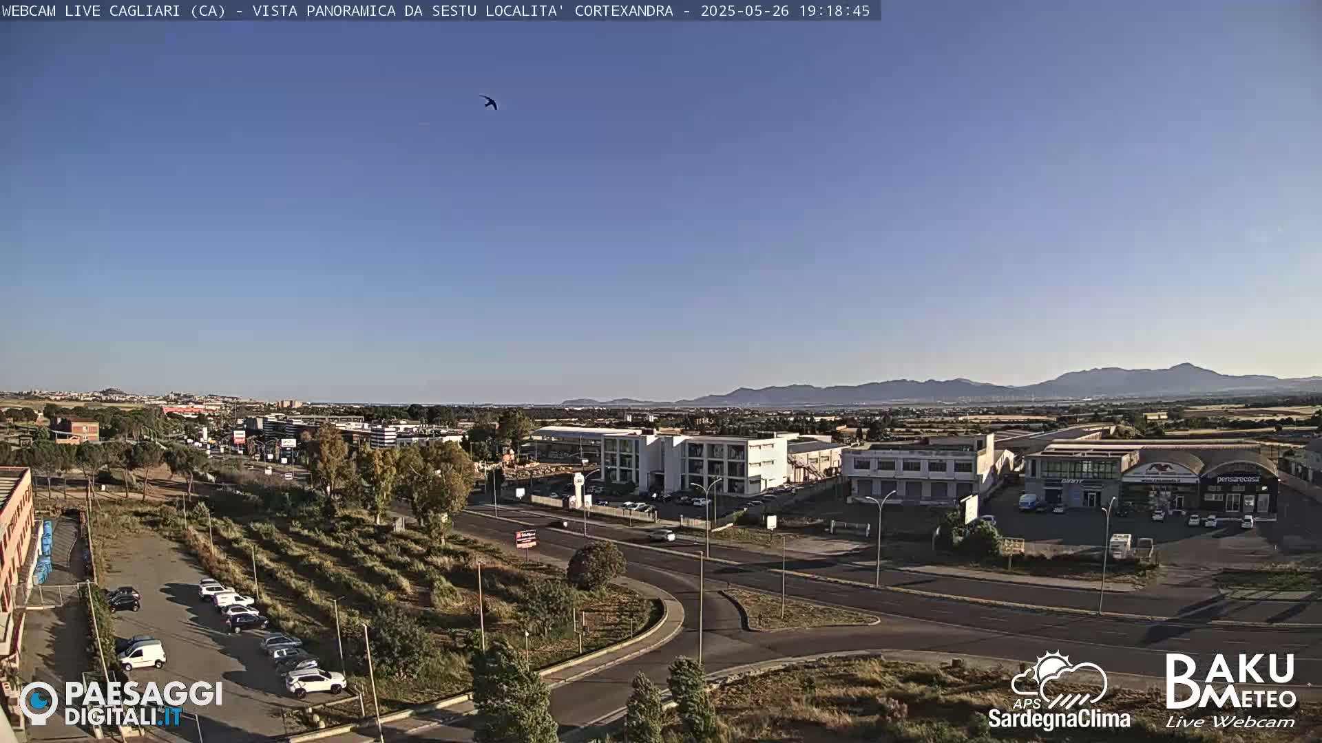 A clear, sunny view of a suburban area with a road, buildings, and parked cars, extending to distant mountains under a bright blue sky.