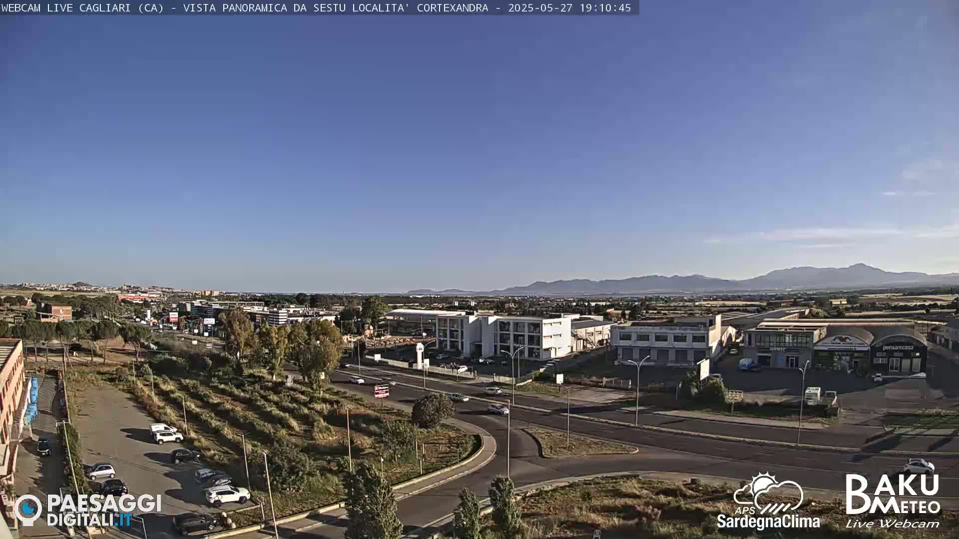 A clear, sunny day reveals a panoramic view of a city's outskirts, featuring roads, buildings, and sparse vegetation, with mountains visible in the distance.