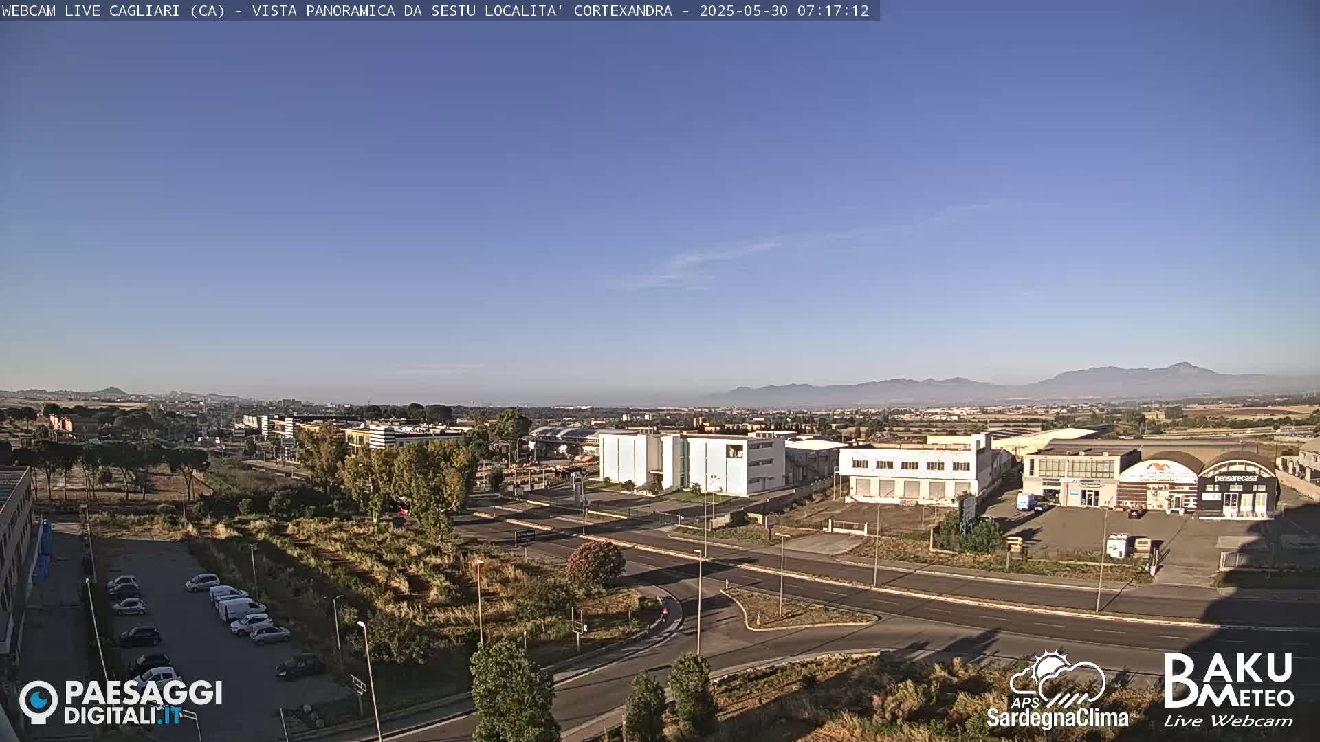 A clear, sunny day reveals a panoramic view of a city with industrial buildings and a parking lot, situated in front of a distant mountain range.