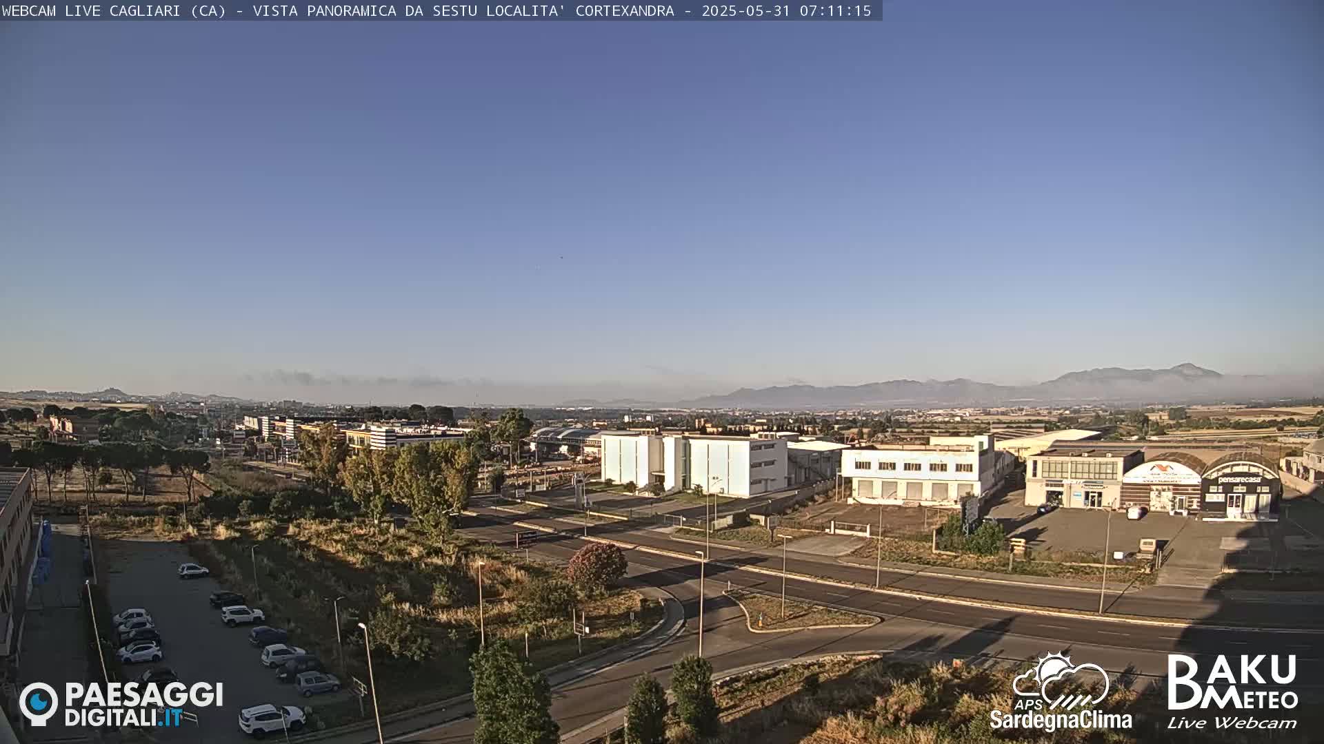 A panoramic view of a city with light industrial buildings and a parking lot under a clear, sunny sky with distant mountains partially obscured by morning mist.