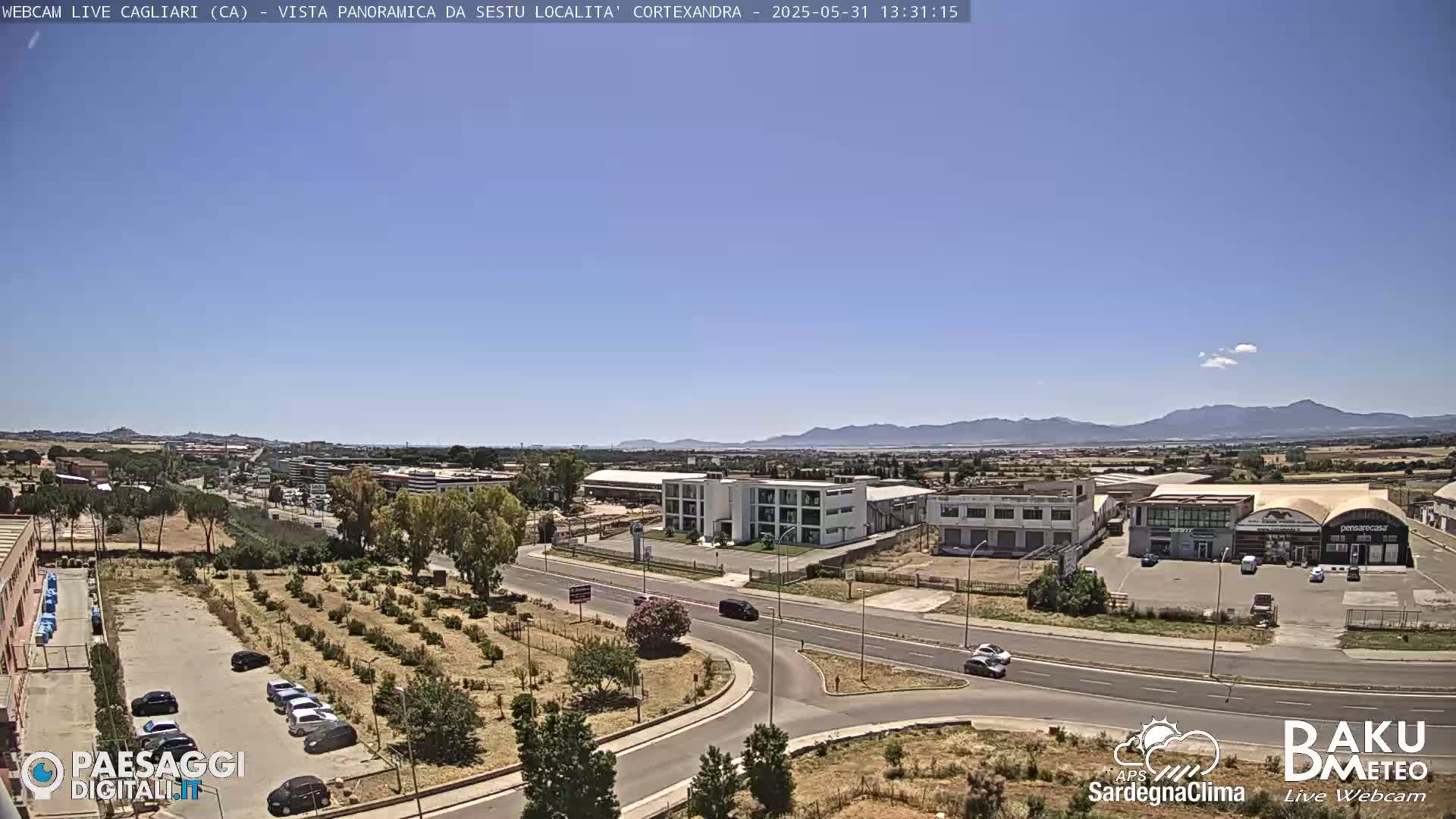 A sunny day reveals a panoramic view of an industrial area and surrounding landscape, featuring a road intersection, buildings, and a few cars parked and driving.