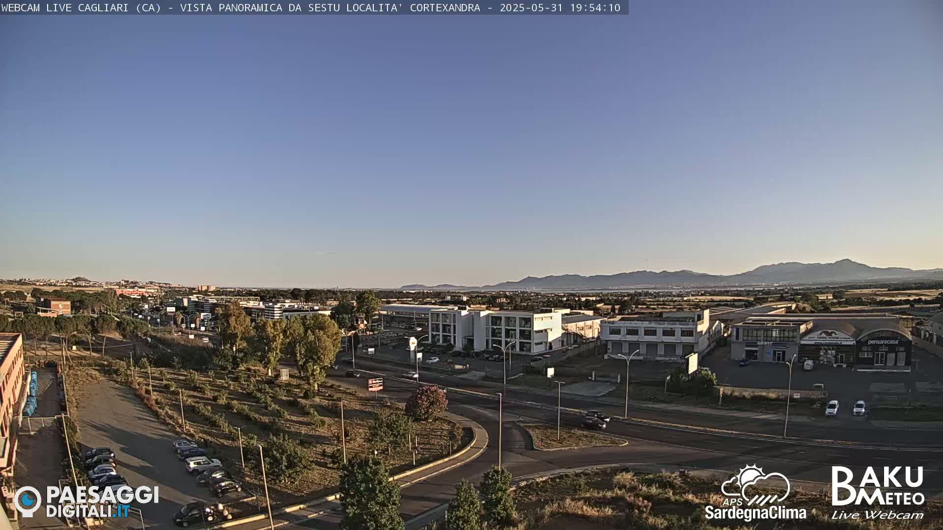 A clear, late afternoon view of a city with low-rise buildings and a road intersection, backed by distant mountains under a mostly clear sky.