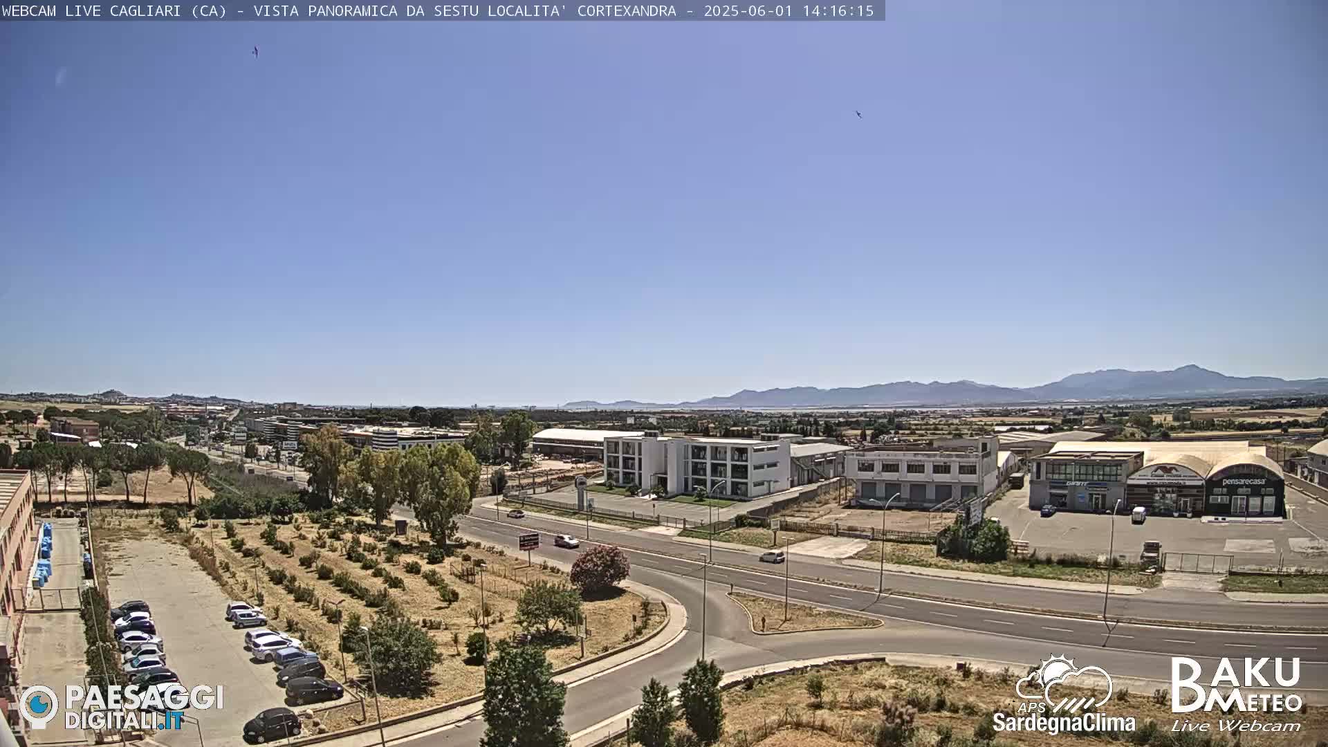 A sunny day reveals a panoramic view of a road system, industrial buildings, and a parking lot surrounded by sparse vegetation, with mountains visible in the distance.