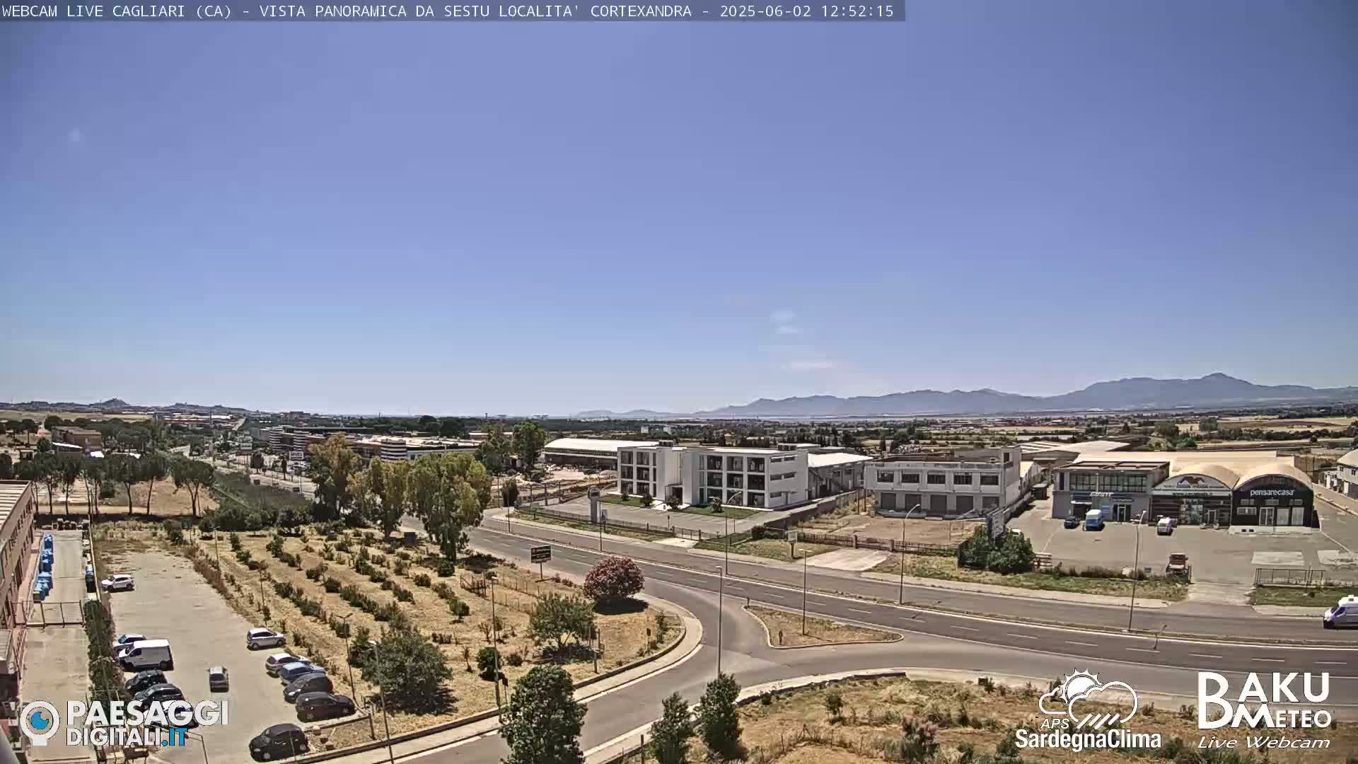 A clear, sunny day overlooks a road intersection, industrial buildings, and a sparse orchard, with mountains visible in the distance.
