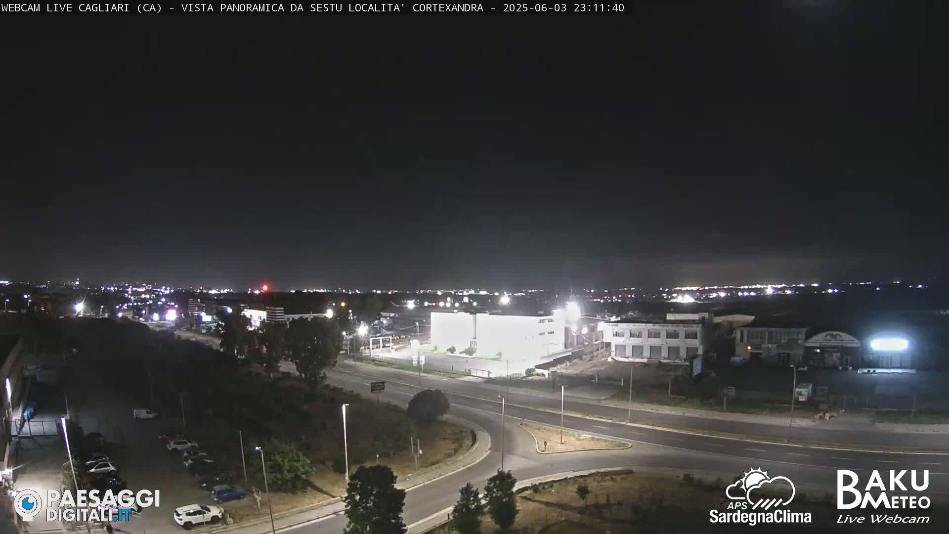 A nighttime, aerial view of a road intersection and surrounding industrial area, with city lights visible in the distance under a clear, dark sky.