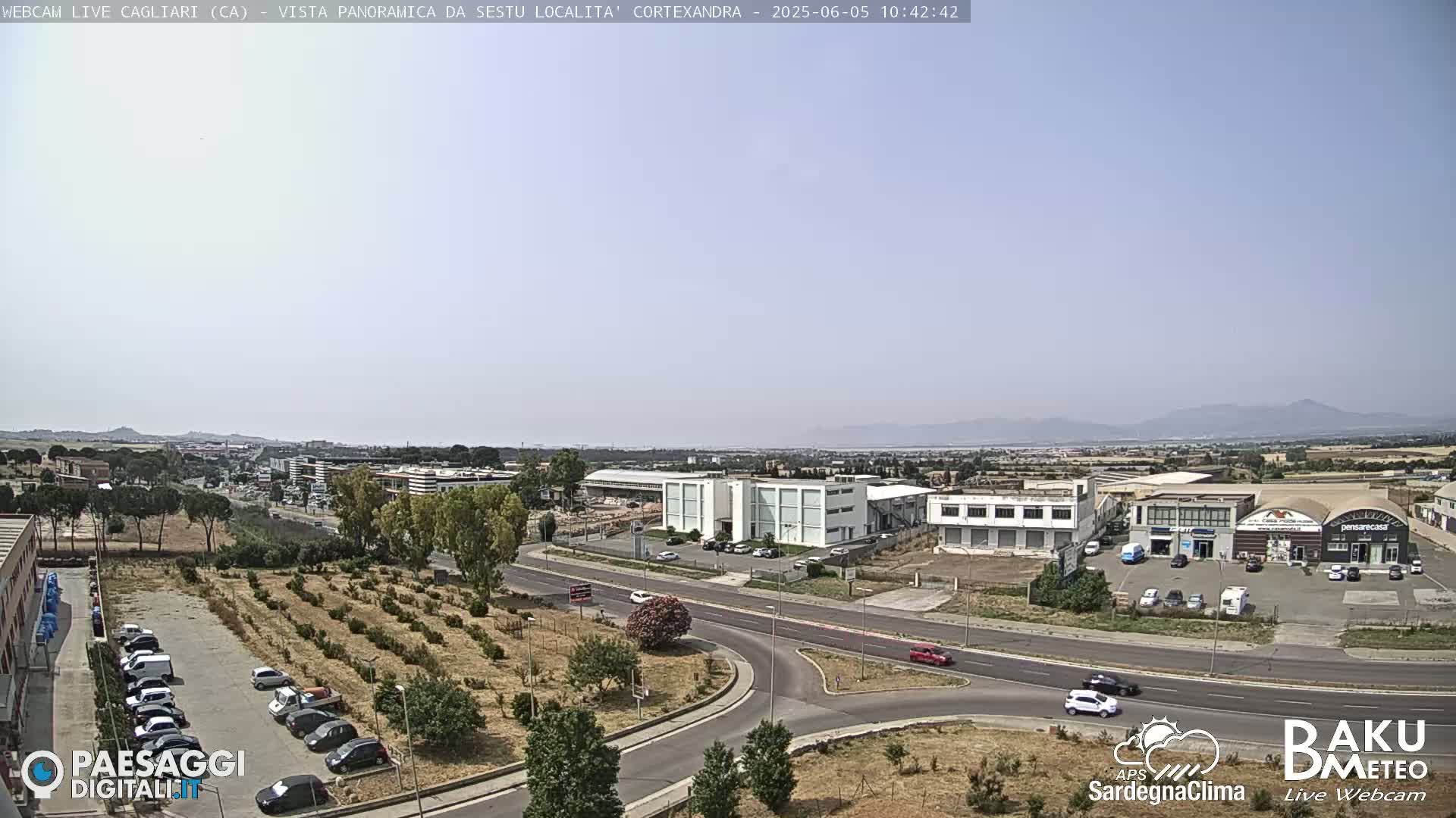 A panoramic view of a city under a clear, sunny sky shows roads, buildings, and a parking lot with cars, surrounded by dry, brown vegetation.