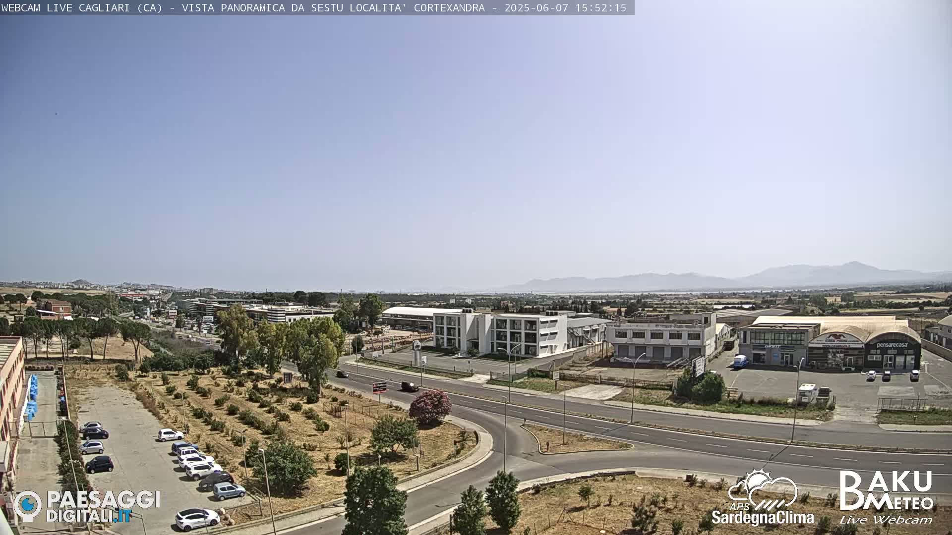 A sunny, high-angle view of a roadway intersection, surrounded by industrial buildings, businesses, and sparse vegetation, with distant mountains visible under a clear sky.