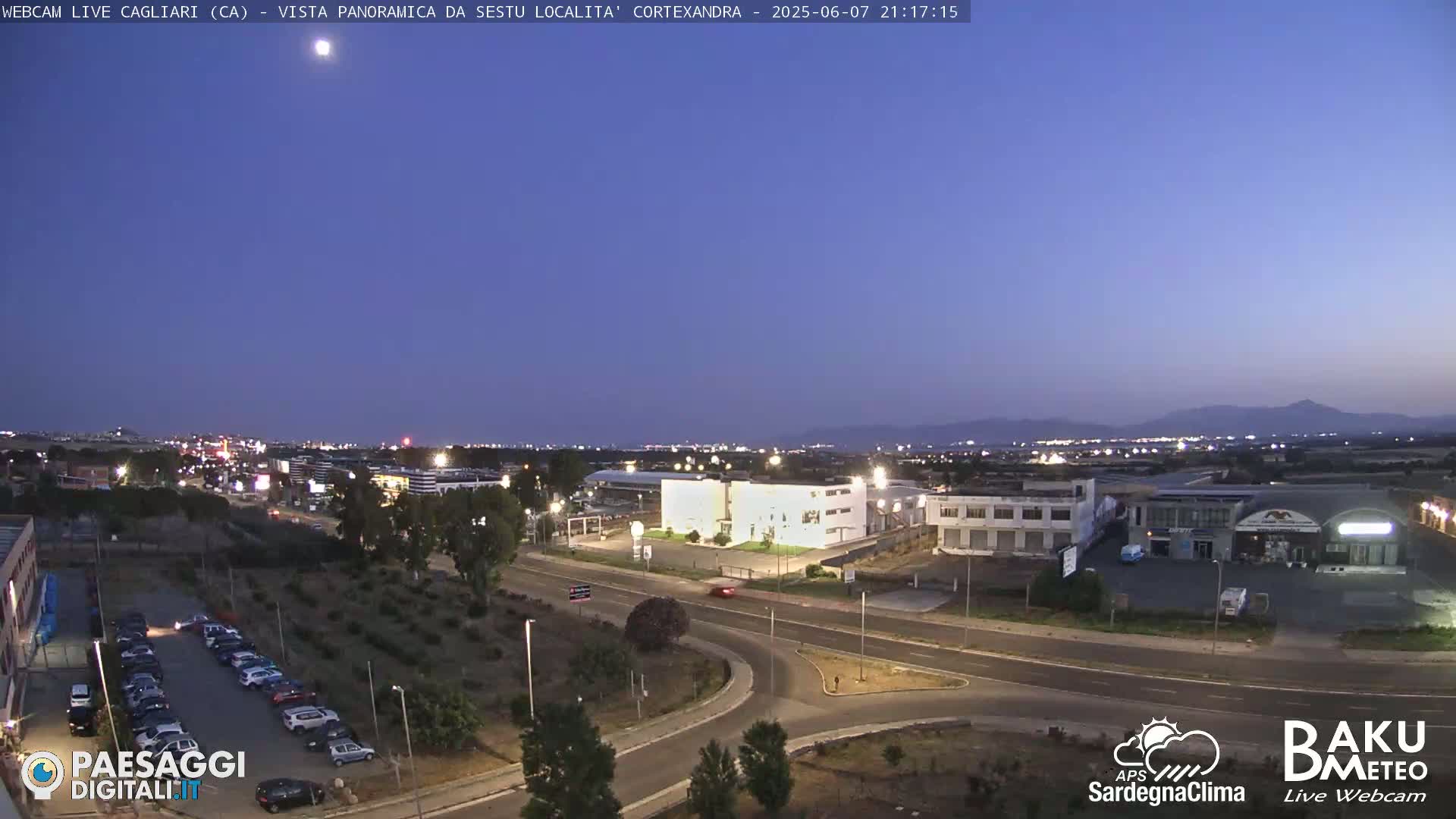 A twilight view of a city at night, showing roads, buildings, and parking lots, with mountains in the distance under a clear sky.