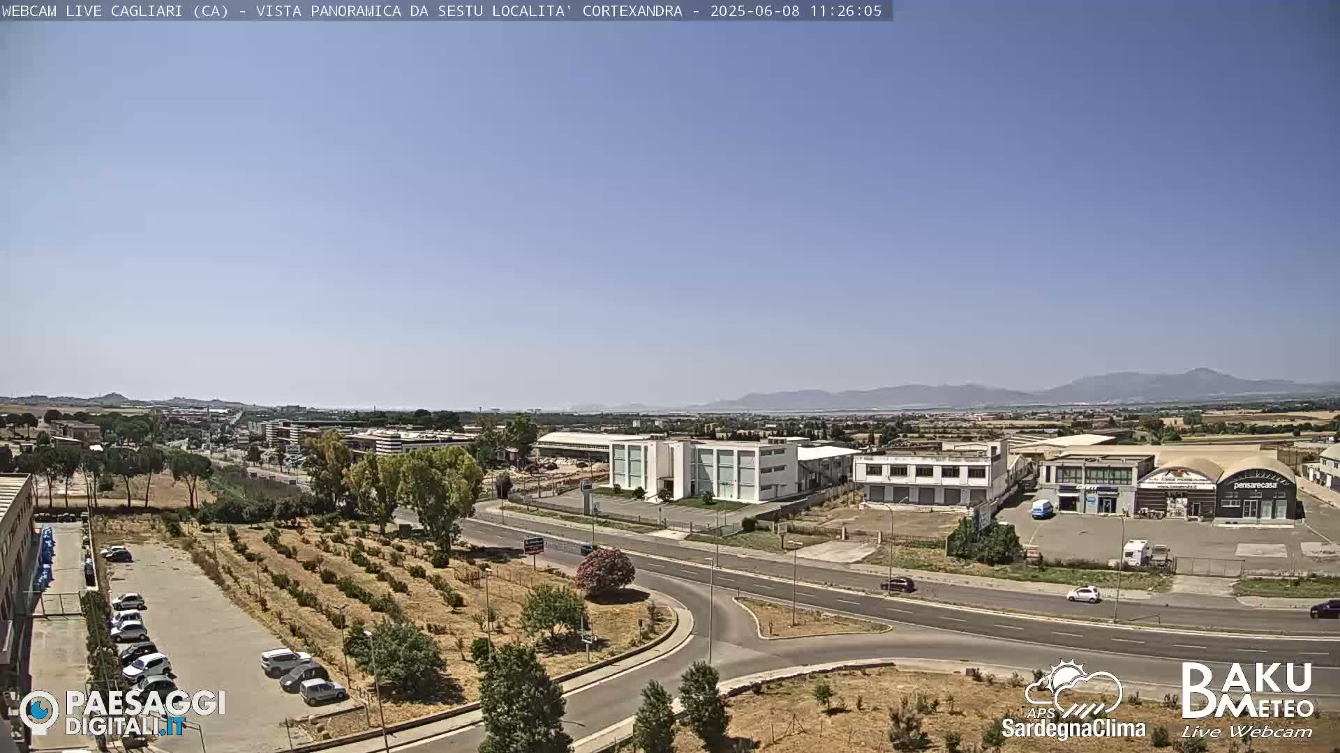 A sunny, clear day reveals a panoramic view of an industrial area near a highway intersection, with mountains in the distance and a parking lot containing several cars.