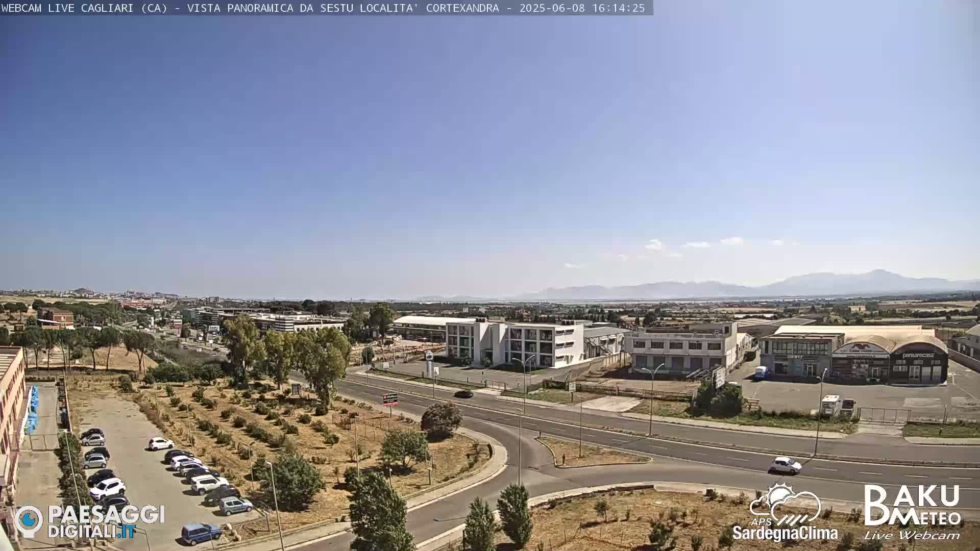 A panoramic view of a city landscape on a clear, sunny day, showing industrial buildings, roads, parked cars, and distant mountains.