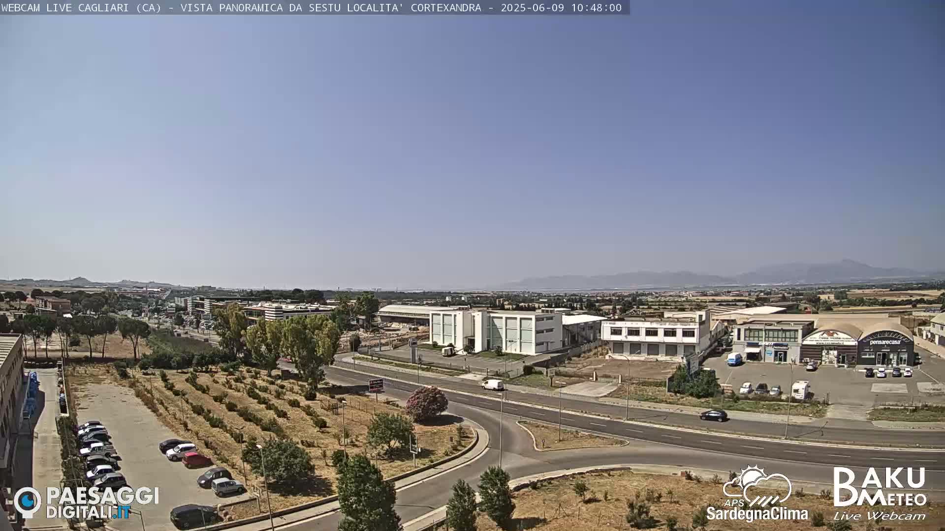 A panoramic view of a city under a clear, sunny sky, showing roads, buildings, and a sparse landscape with some vegetation.